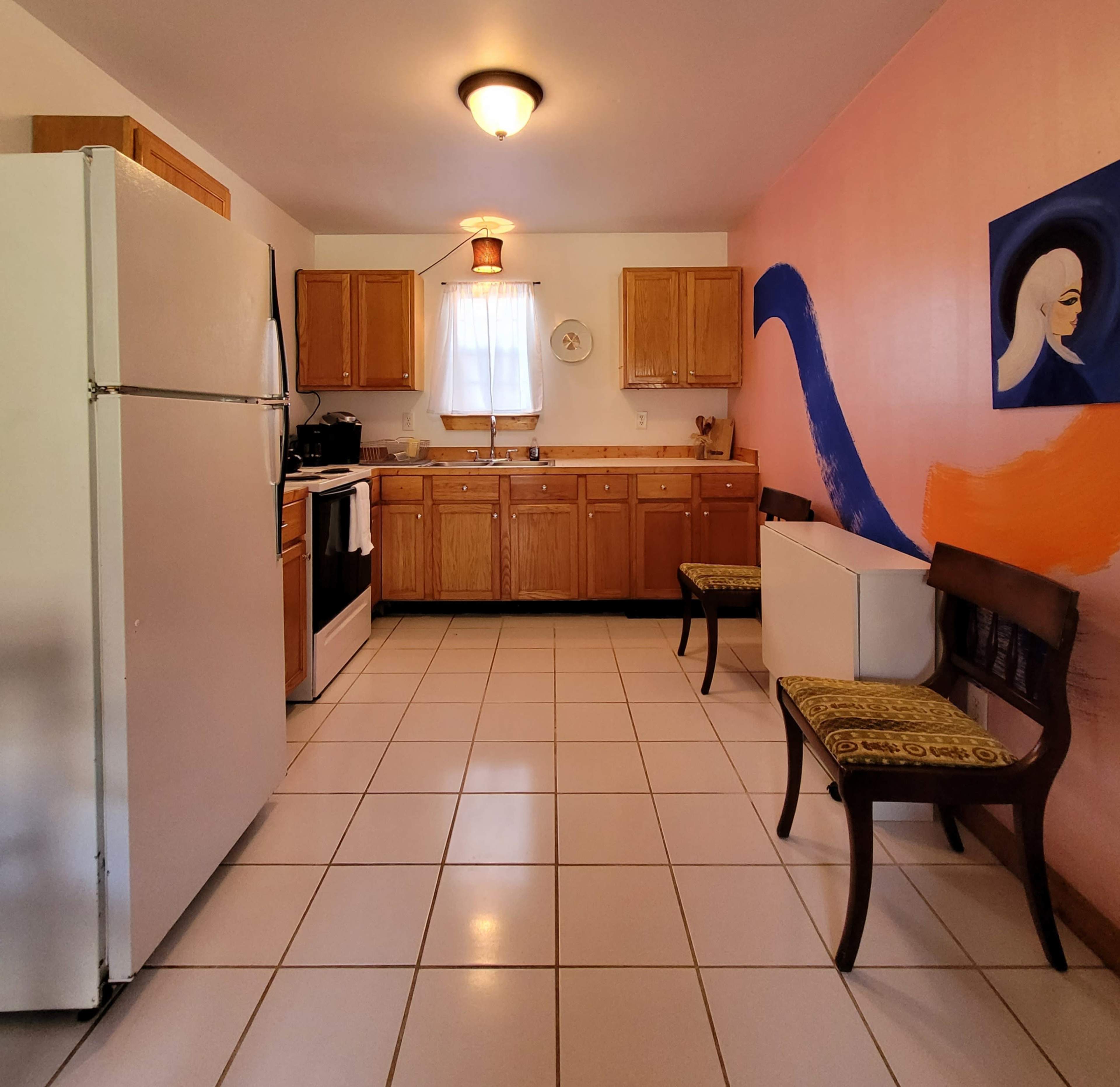 The kitchen features wooden cabinets, a white refrigerator, and two chairs against a pink wall decorated with abstract art.