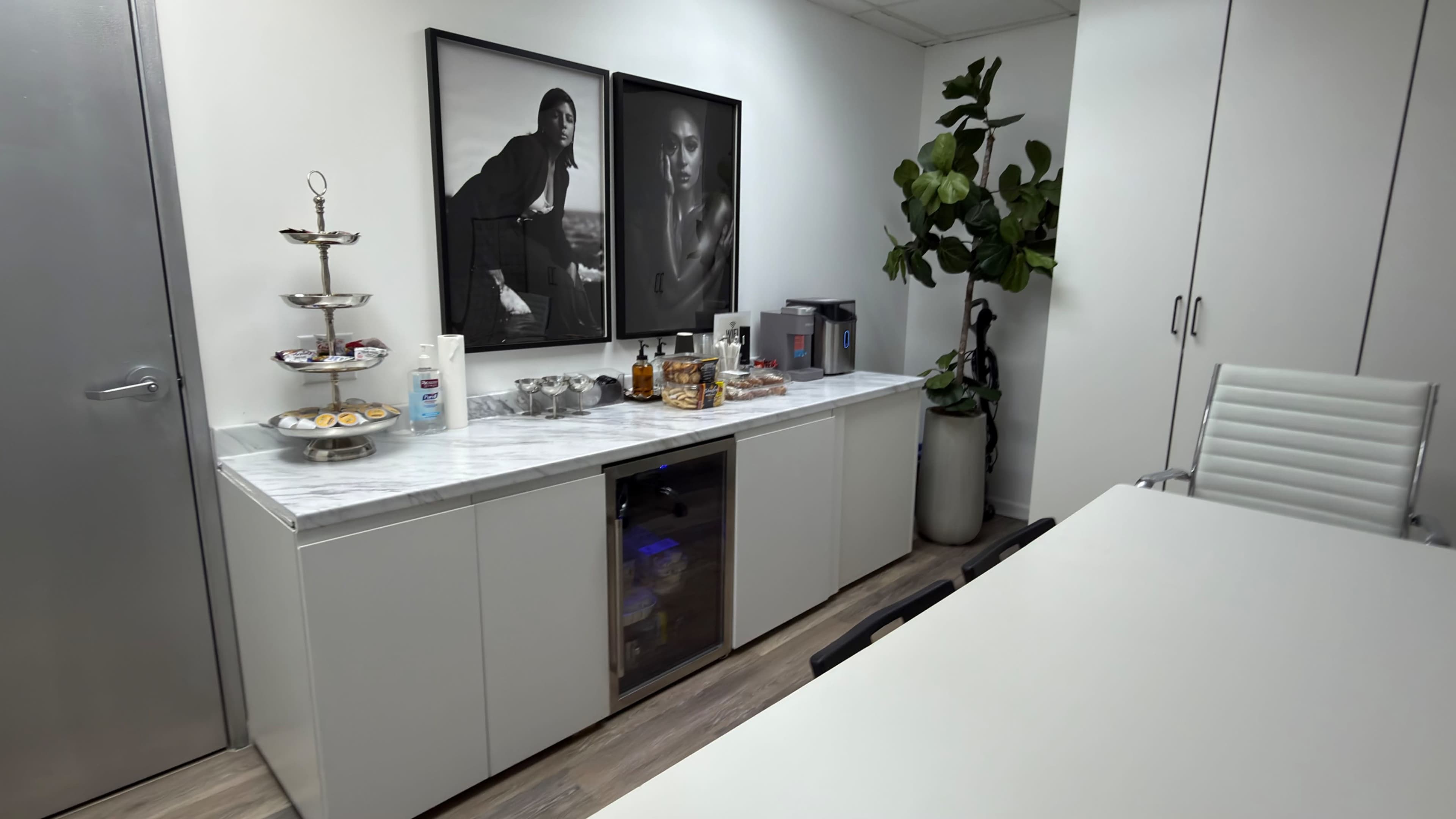 The image shows a modern office kitchenette with a marble countertop, a tiered serving stand with pastries, a mini bar with alcoholic beverages, two framed black-and-white photographs on the wall, and a large potted plant.