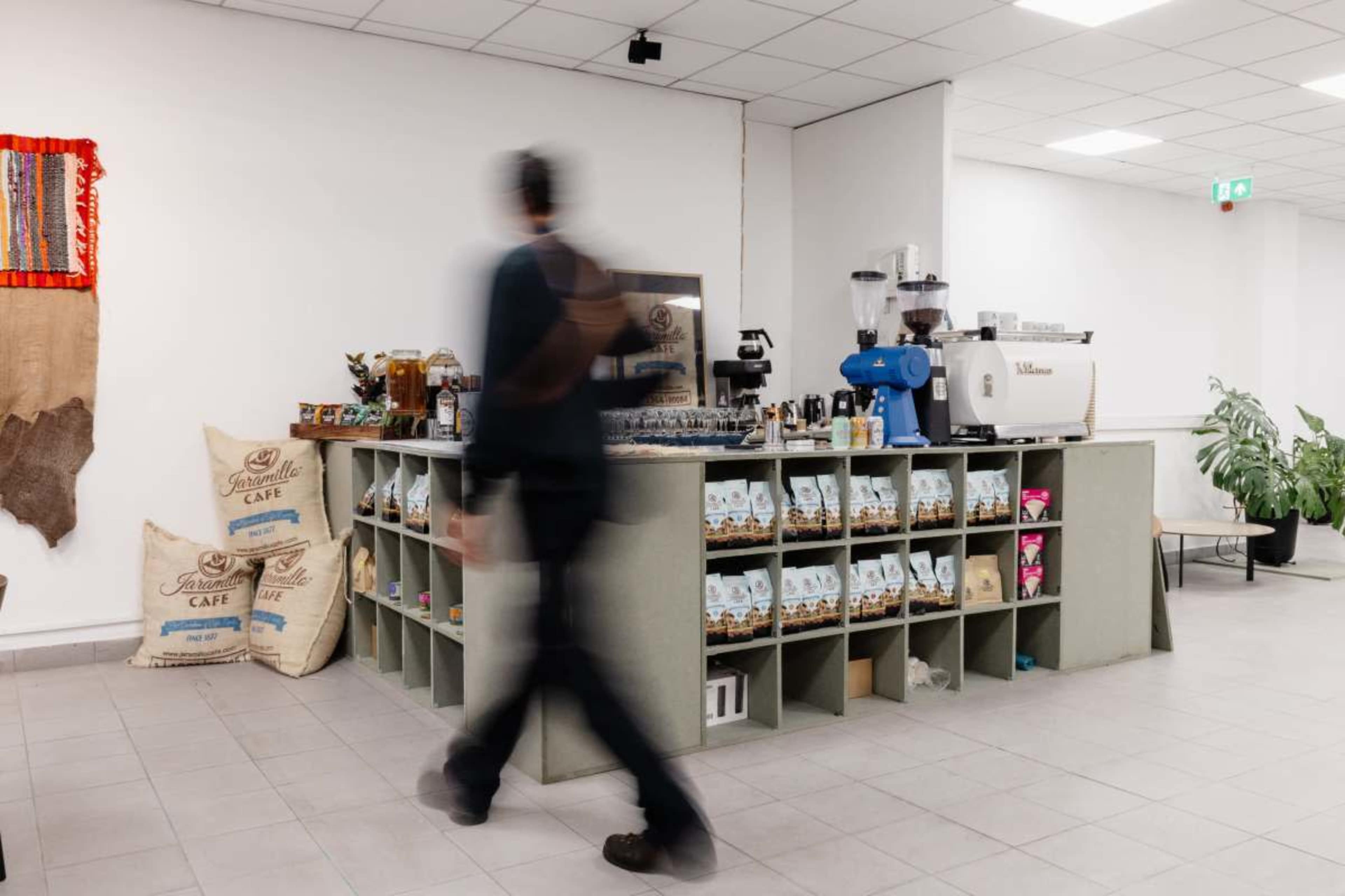 A person walks past a coffee counter displaying bags of coffee and equipment in a bright, modern cafe.