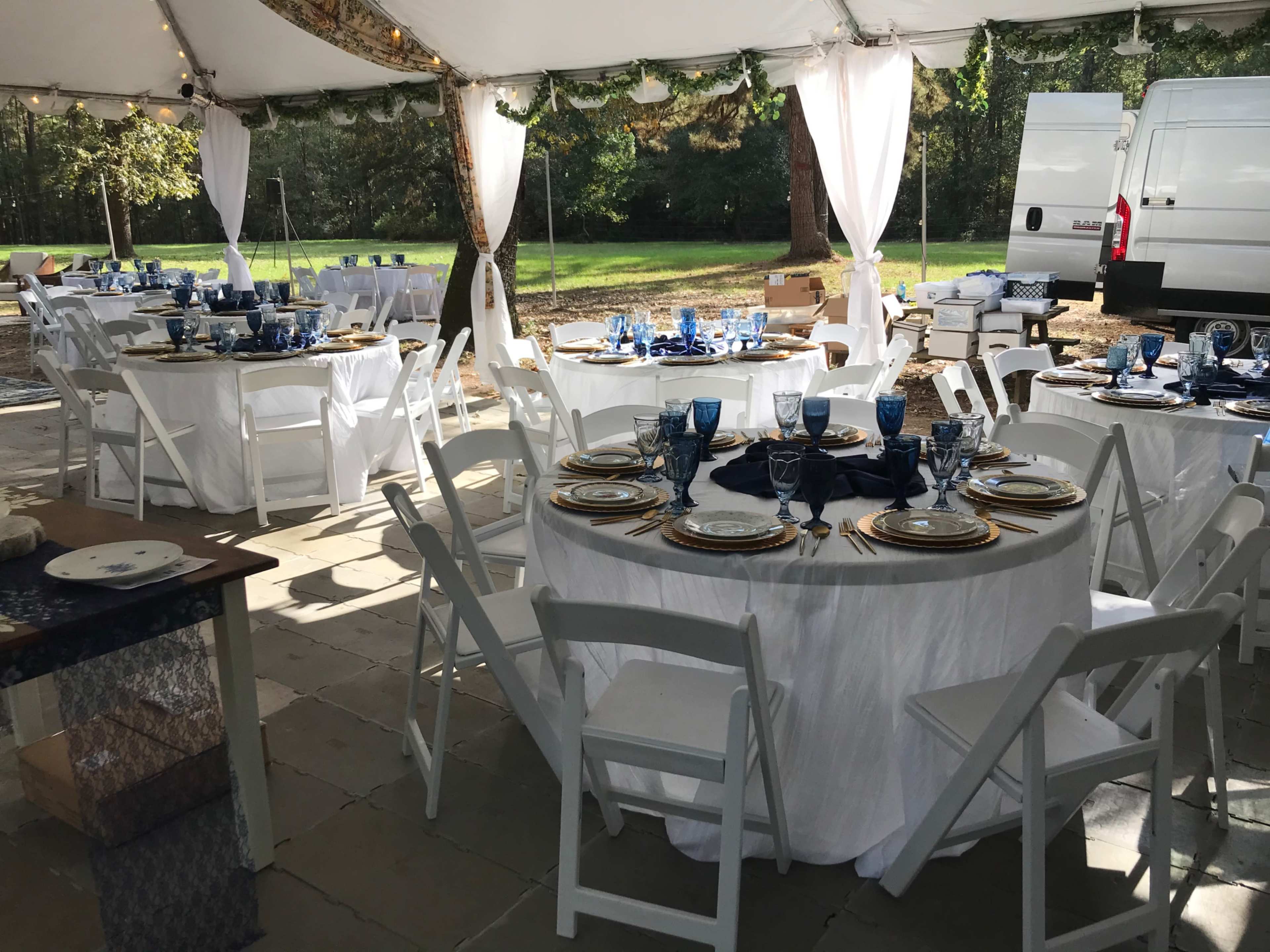 A large white tent is set up for an event, featuring multiple round tables dressed in blue and white tableware, surrounded by folding chairs.
