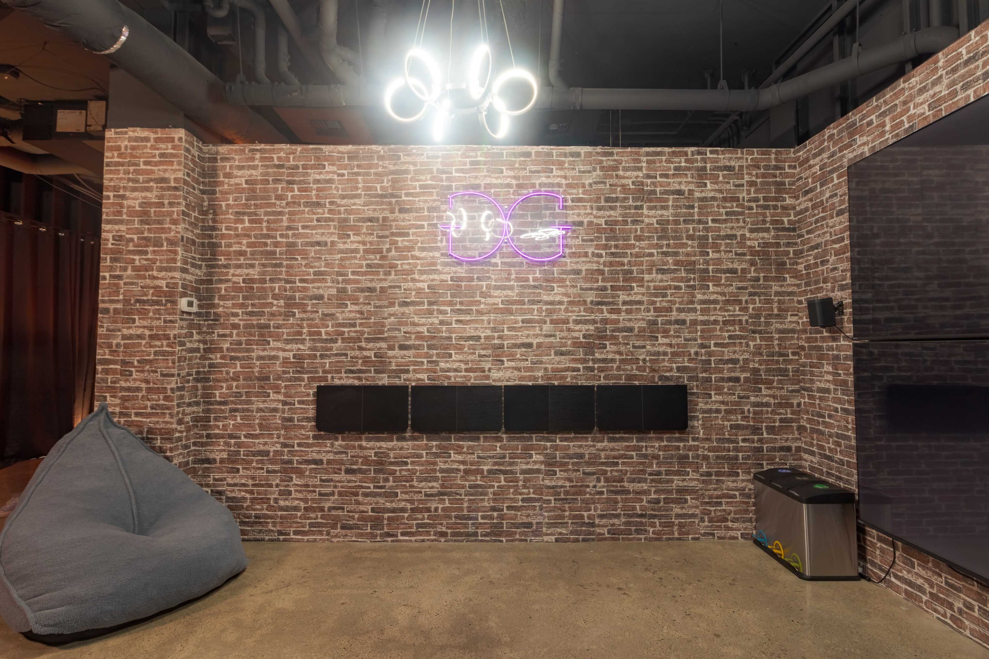 The image shows a brick wall with a neon sign, a row of black panels, a bean bag chair, and a small refrigerator beneath a light fixture in a modern interior space.