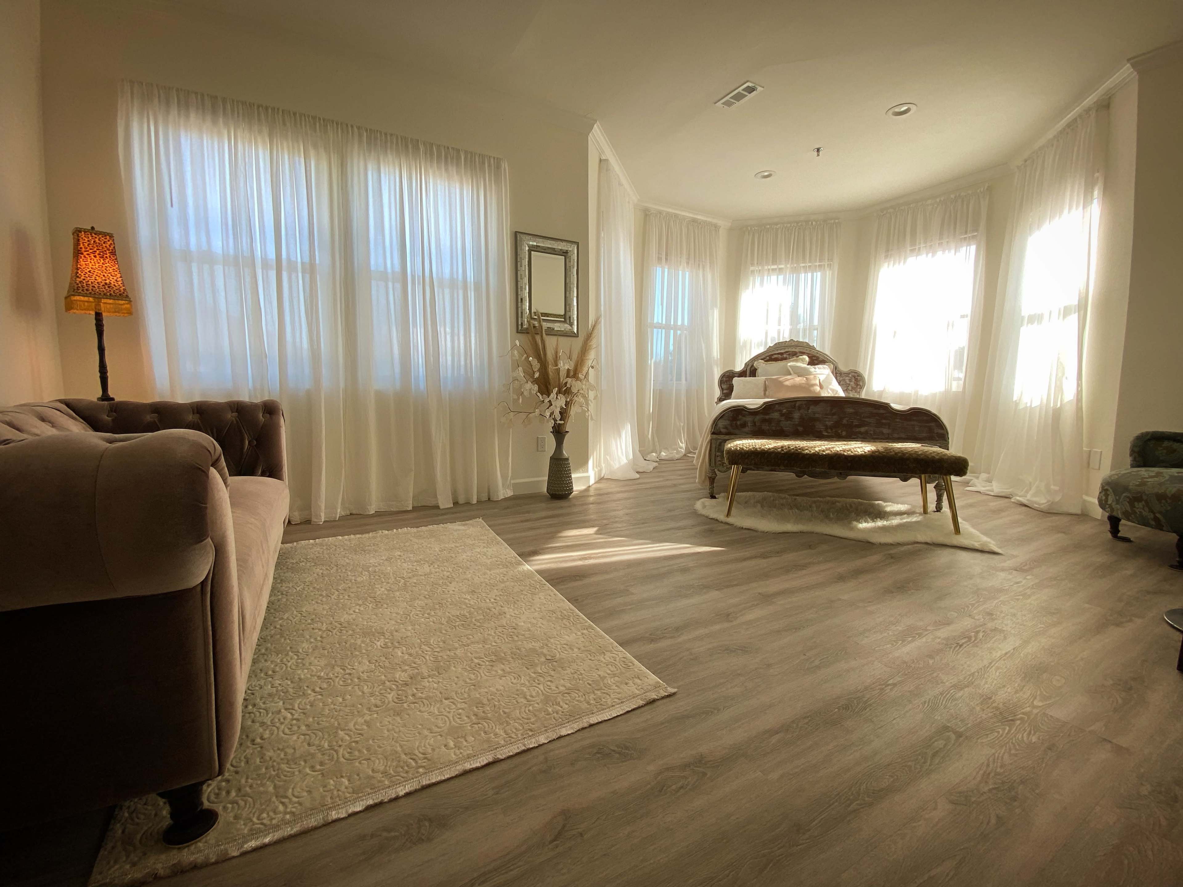 The room features a bed with an ornate headboard, a plush sofa, and large windows draped with sheer curtains that allow natural light to fill the space.