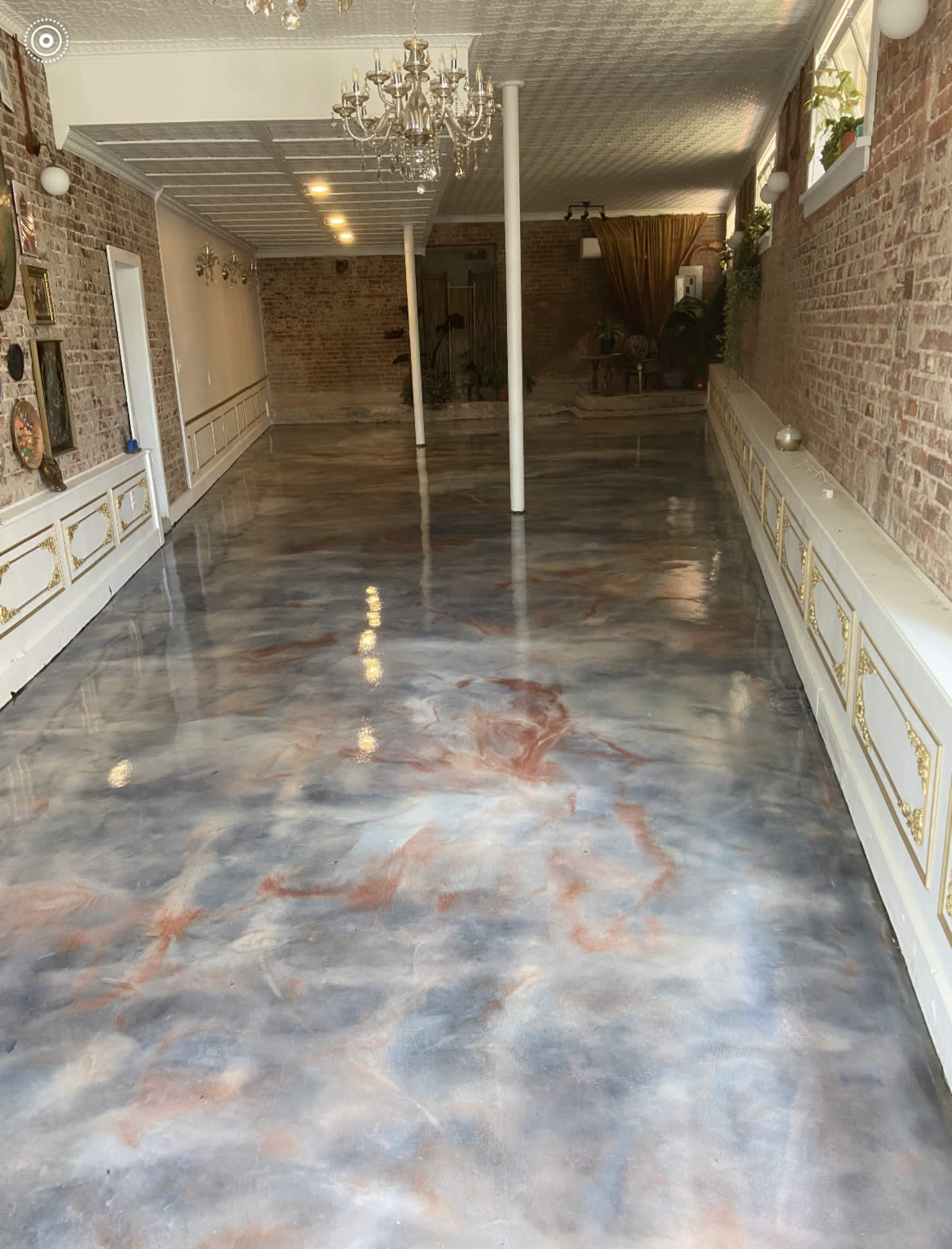 A spacious room with a polished floor featuring a marbled pattern in shades of gray, blue, and rust, flanked by brick walls and decorative chandeliers.