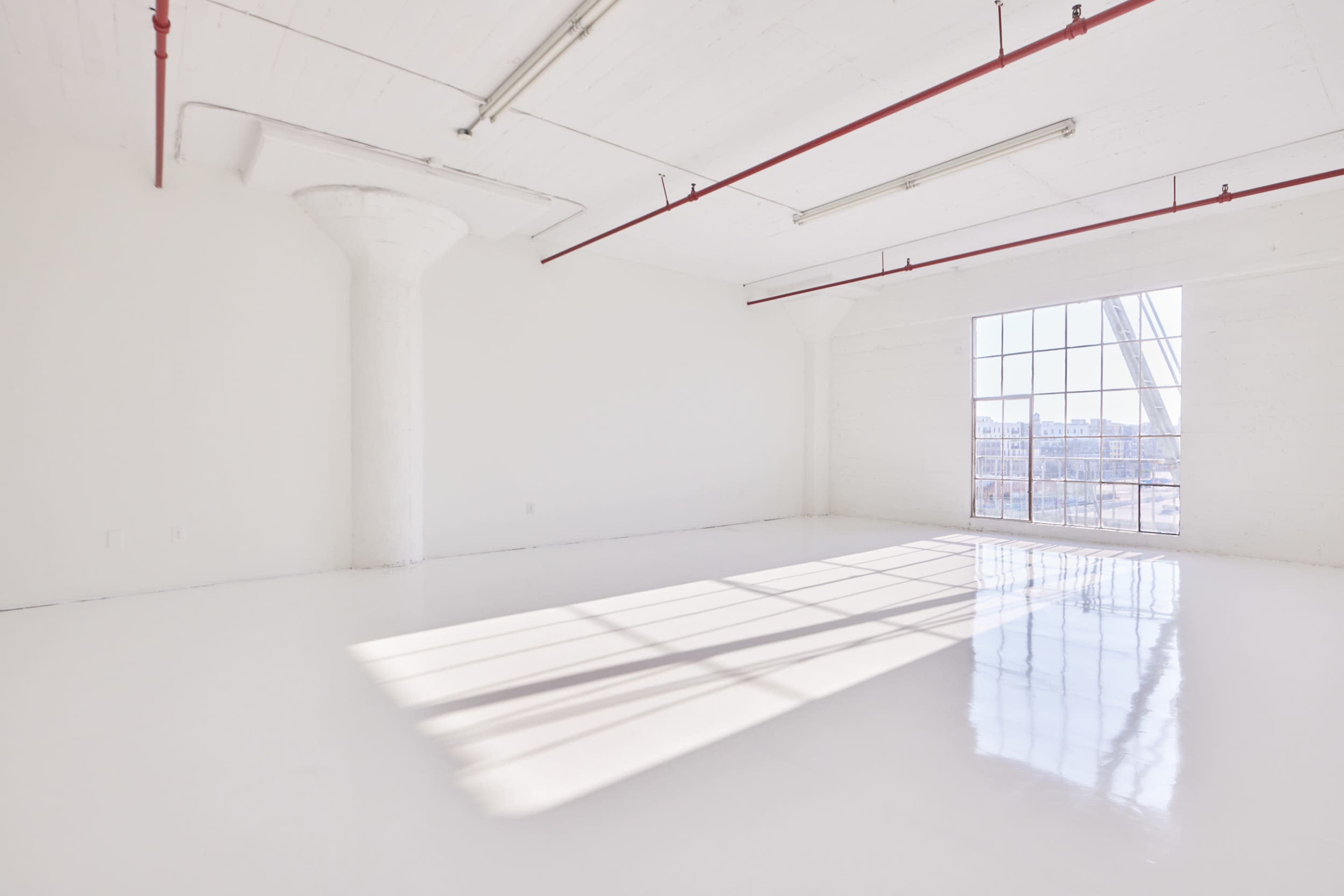 A spacious, white-walled room features large windows that allow sunlight to create shadows on the glossy floor.