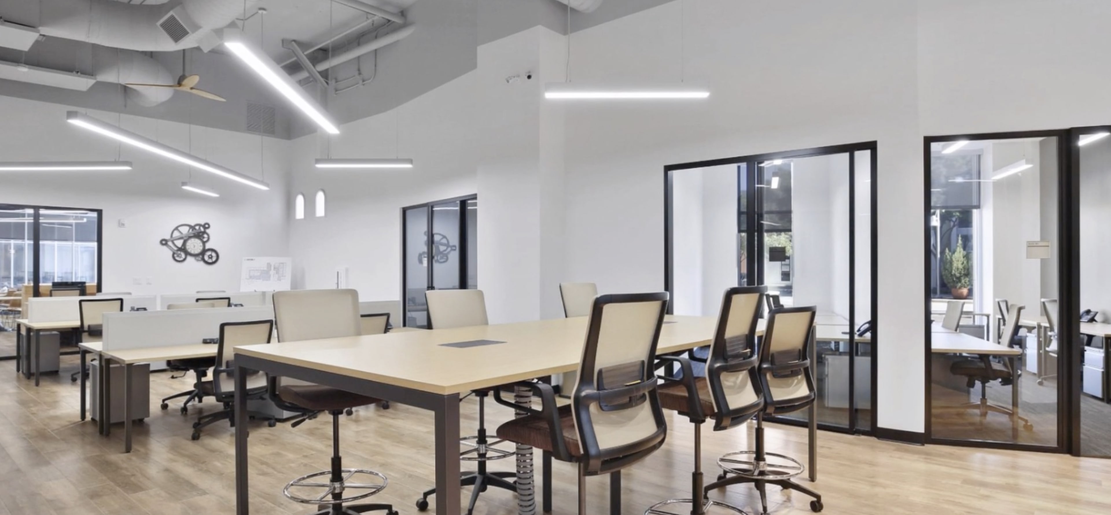 A modern office space with several large tables and ergonomic chairs arranged neatly under bright, linear ceiling lights.