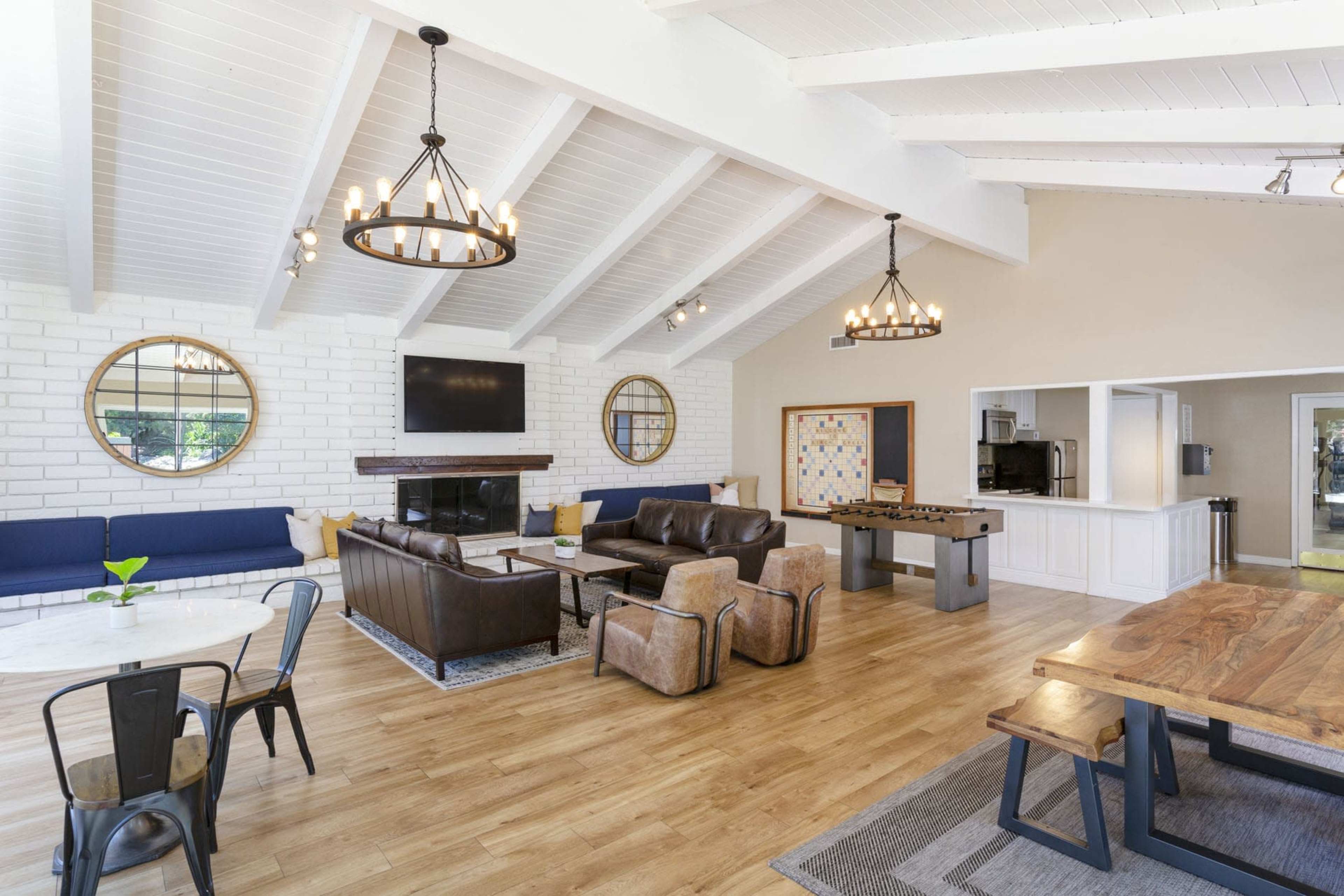 The image shows a spacious lounge area featuring a combination of leather sofas, wooden dining tables, and a brick wall with large mirrors, all under a high ceiling with exposed beams.