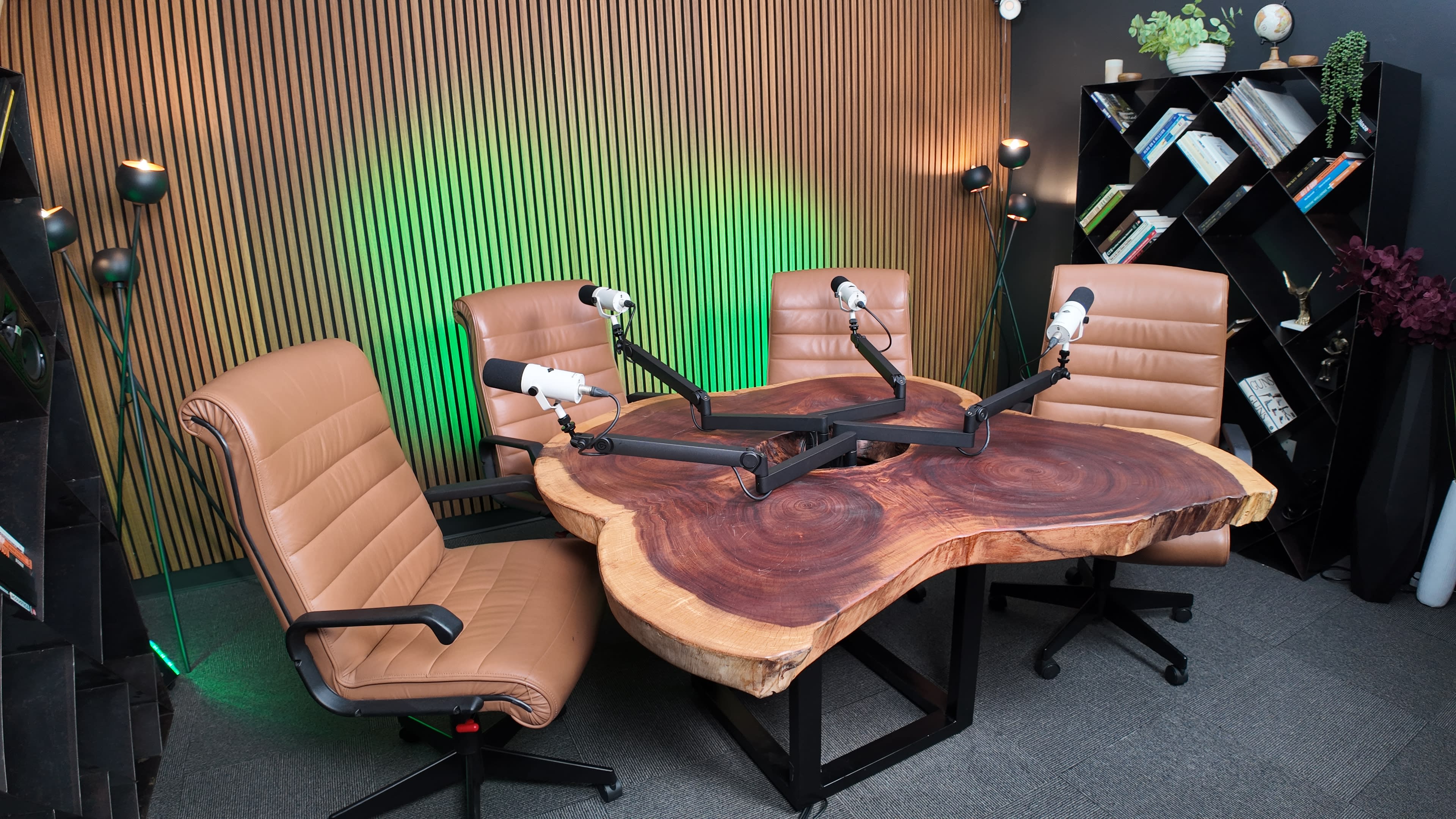 The image shows a podcast recording studio featuring a large wooden table with mounted microphones and four brown leather chairs arranged around it.