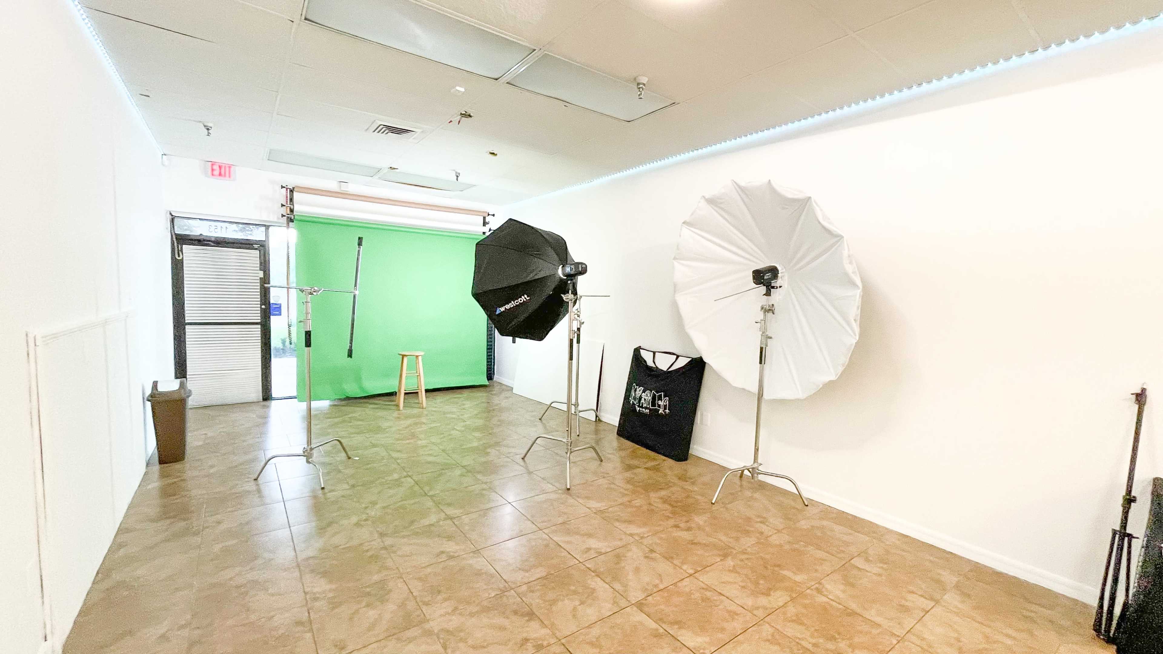 A photography studio featuring two light stands with umbrellas, a green backdrop, a stool, and a bag on the floor.