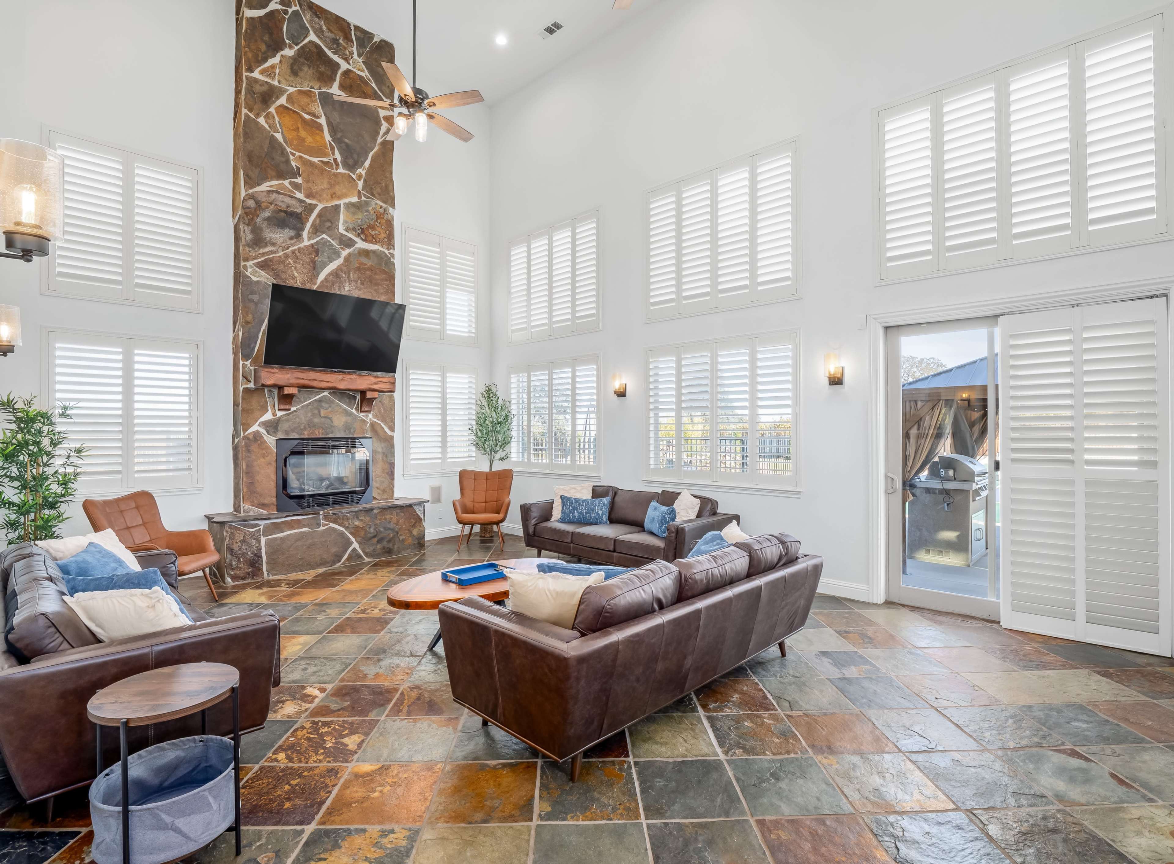 A spacious living room with a stone fireplace, large windows with plantation shutters, and leather sofas arranged around a circular coffee table.