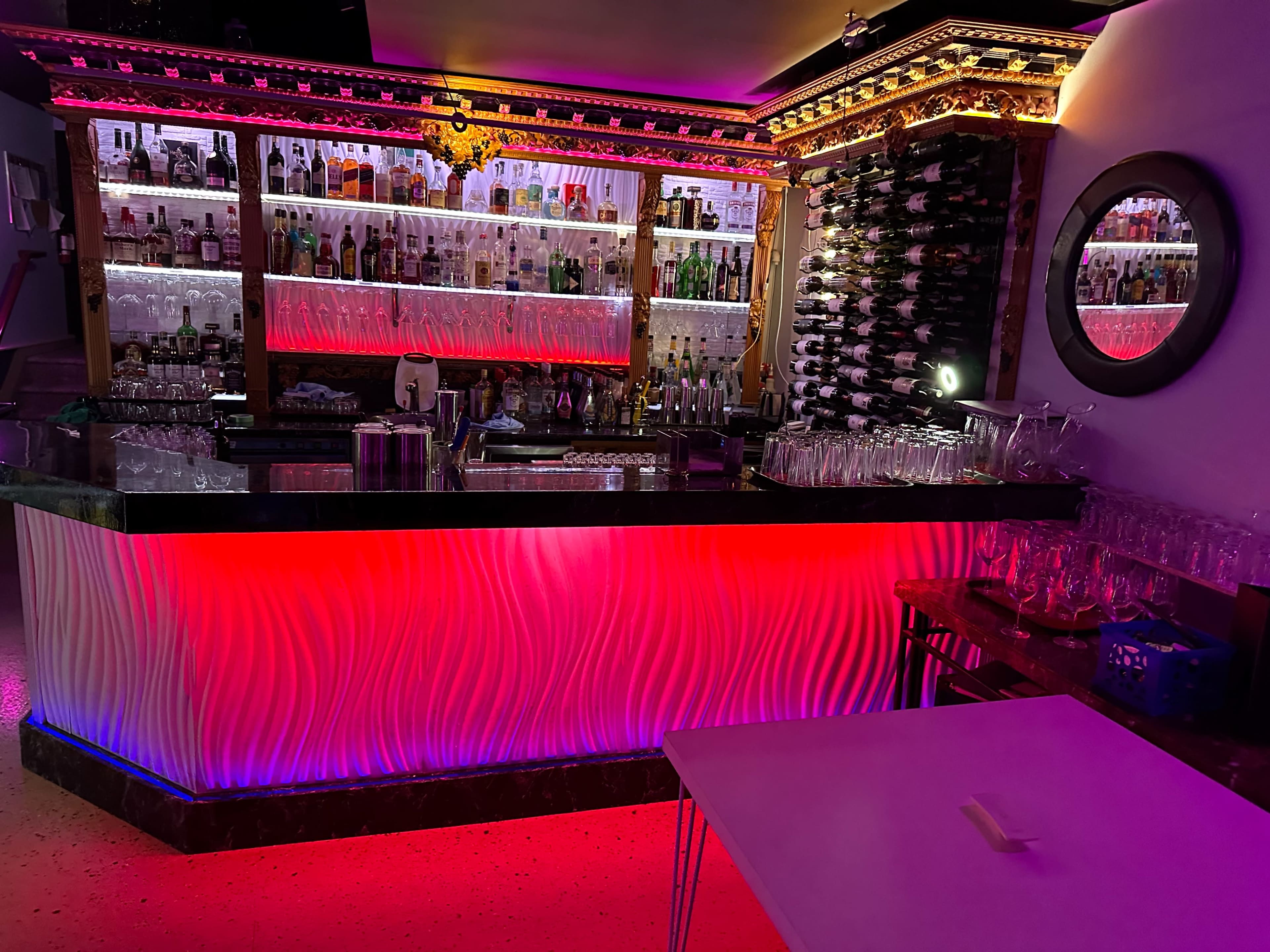 A bar area with a brightly lit counter featuring wave-like designs, surrounded by shelves stocked with various bottles and illuminated by colorful lights.