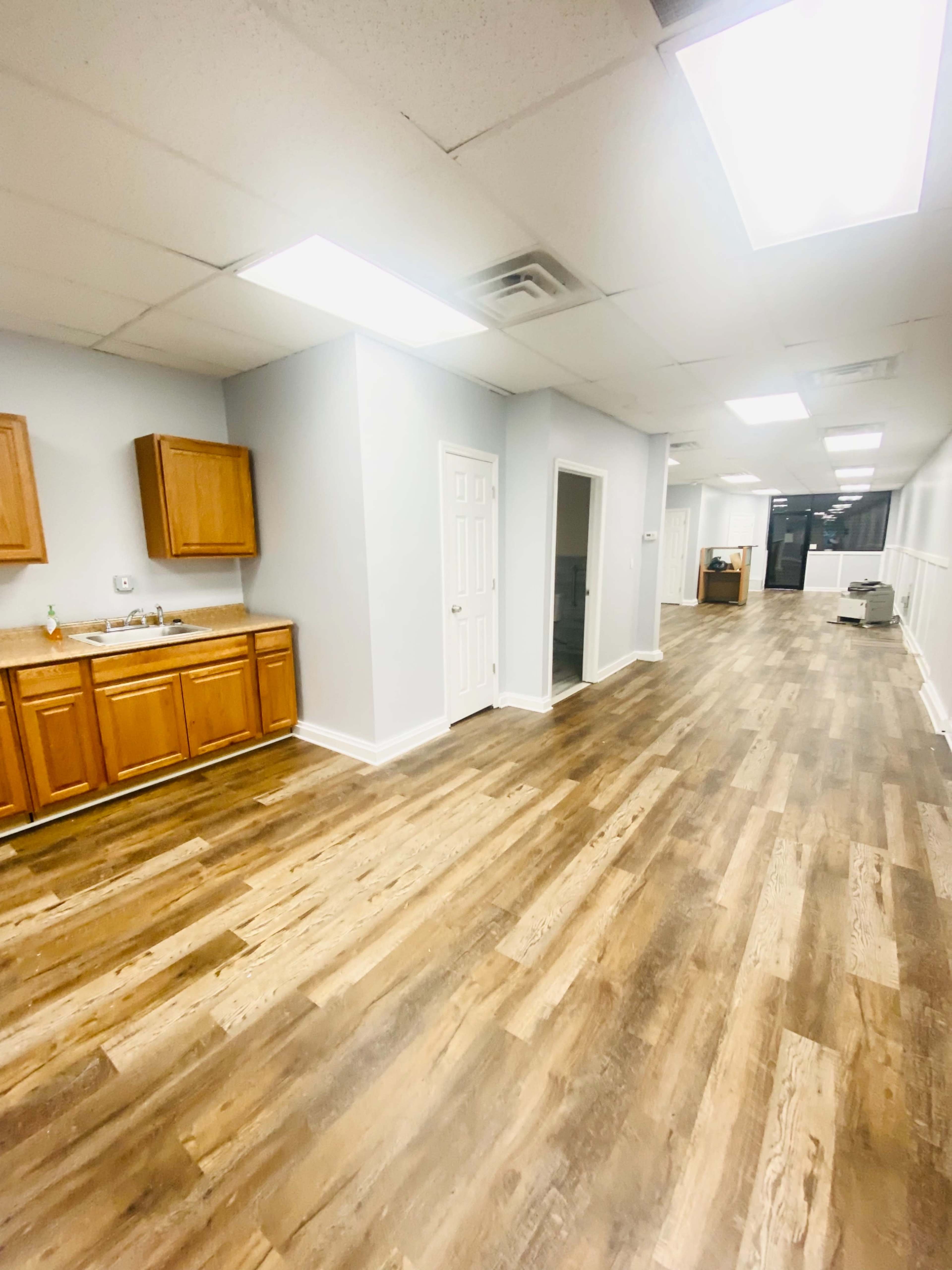 A long hallway in a commercial space with wooden flooring, light gray walls, and a kitchenette area on the left.