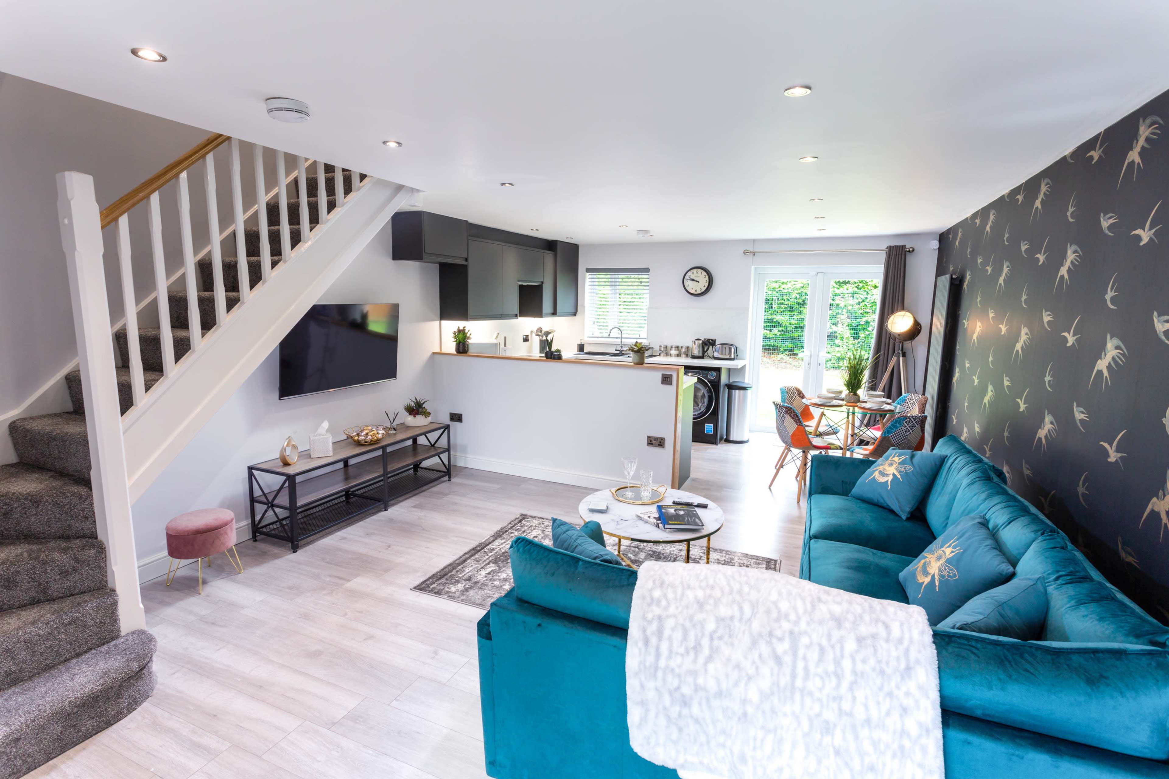 The room features an open-concept living area with a teal sofa, dining space, and a kitchen visible in the background.