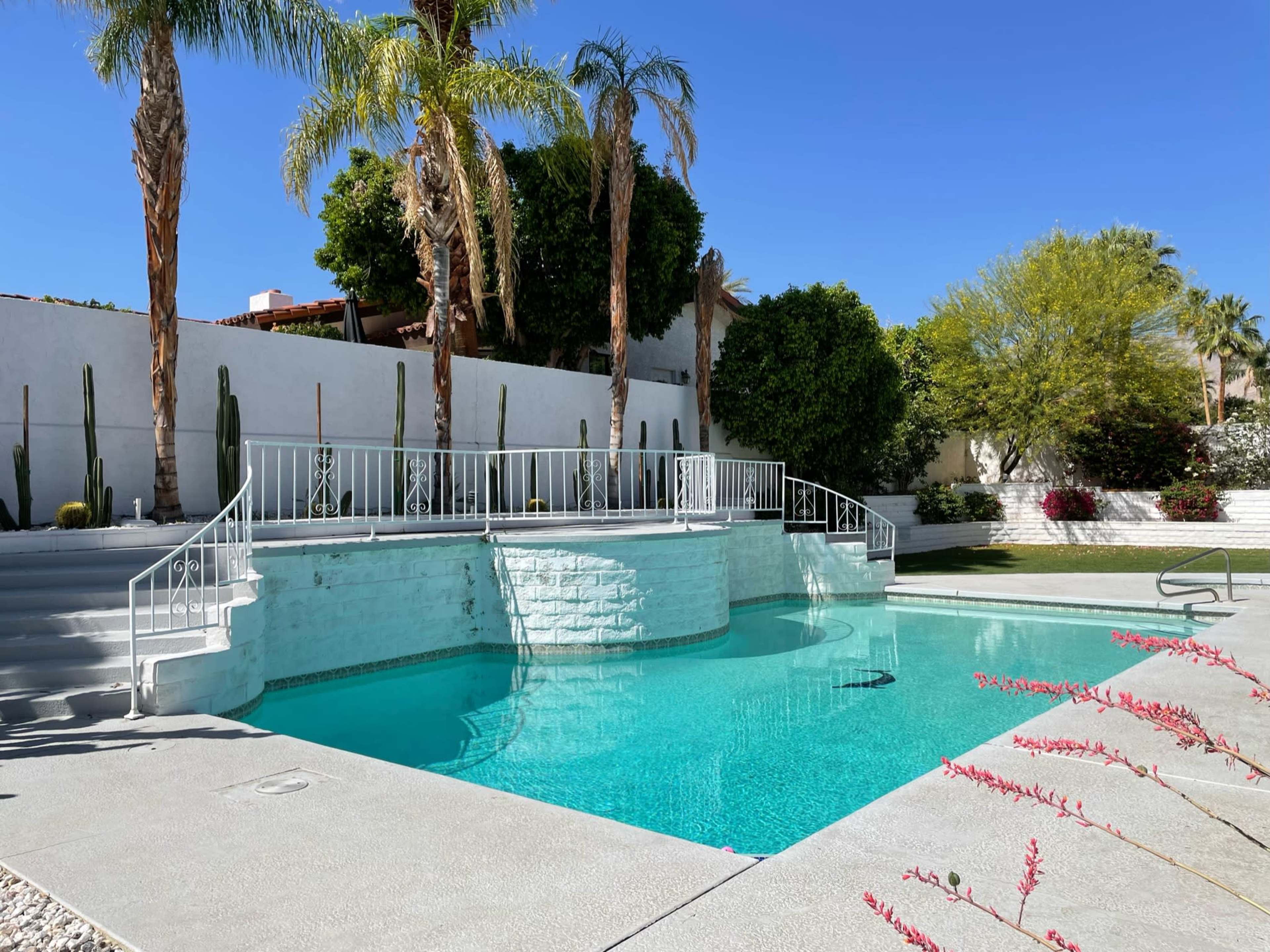 The image shows a landscaped pool area with a turquoise swimming pool, surrounded by palm trees and desert plants, and a white staircase leading up to a garden space.