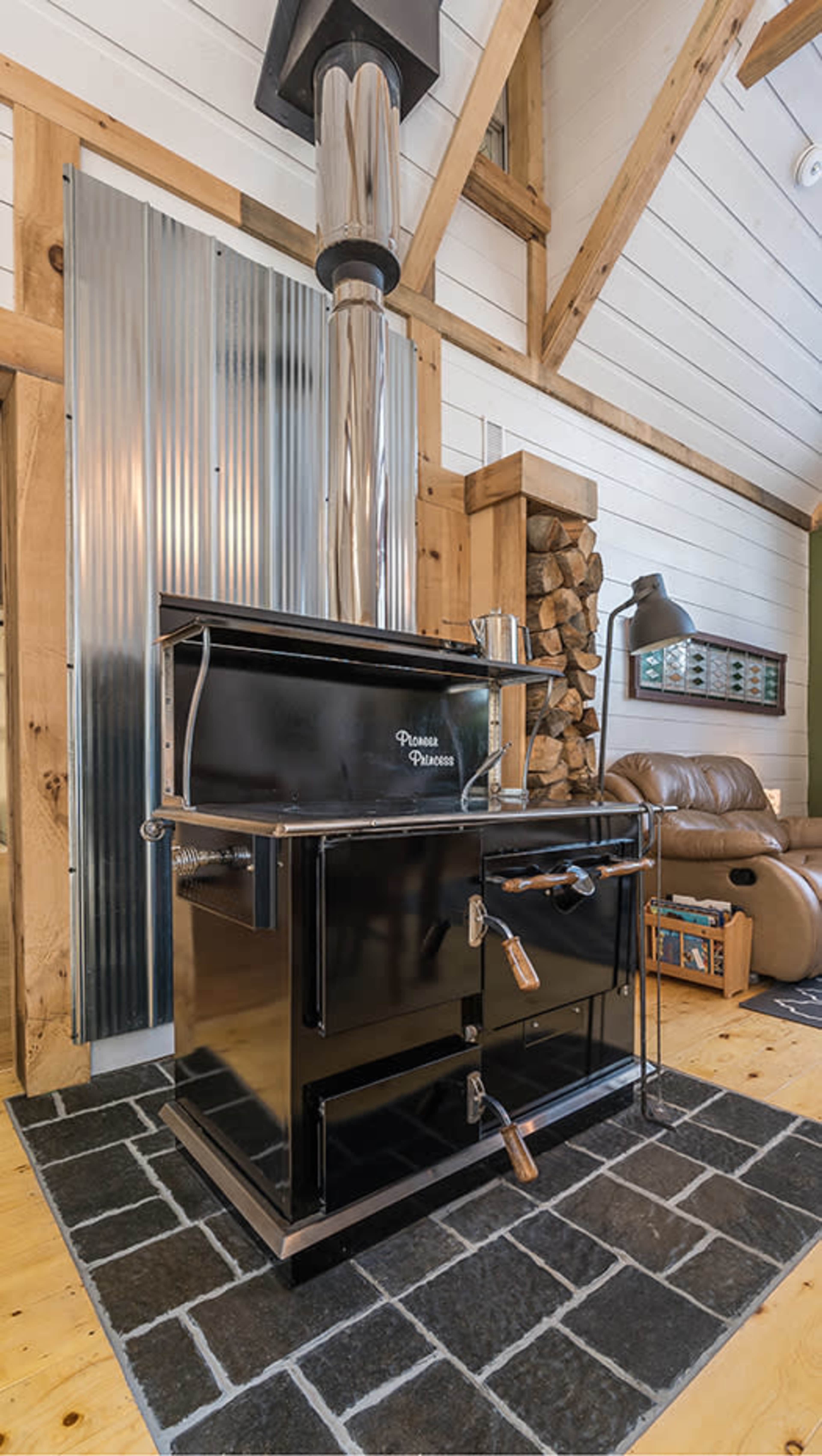 A black cast iron wood-burning stove is positioned on a dark stone floor with logs stacked nearby in a rustic interior setting.