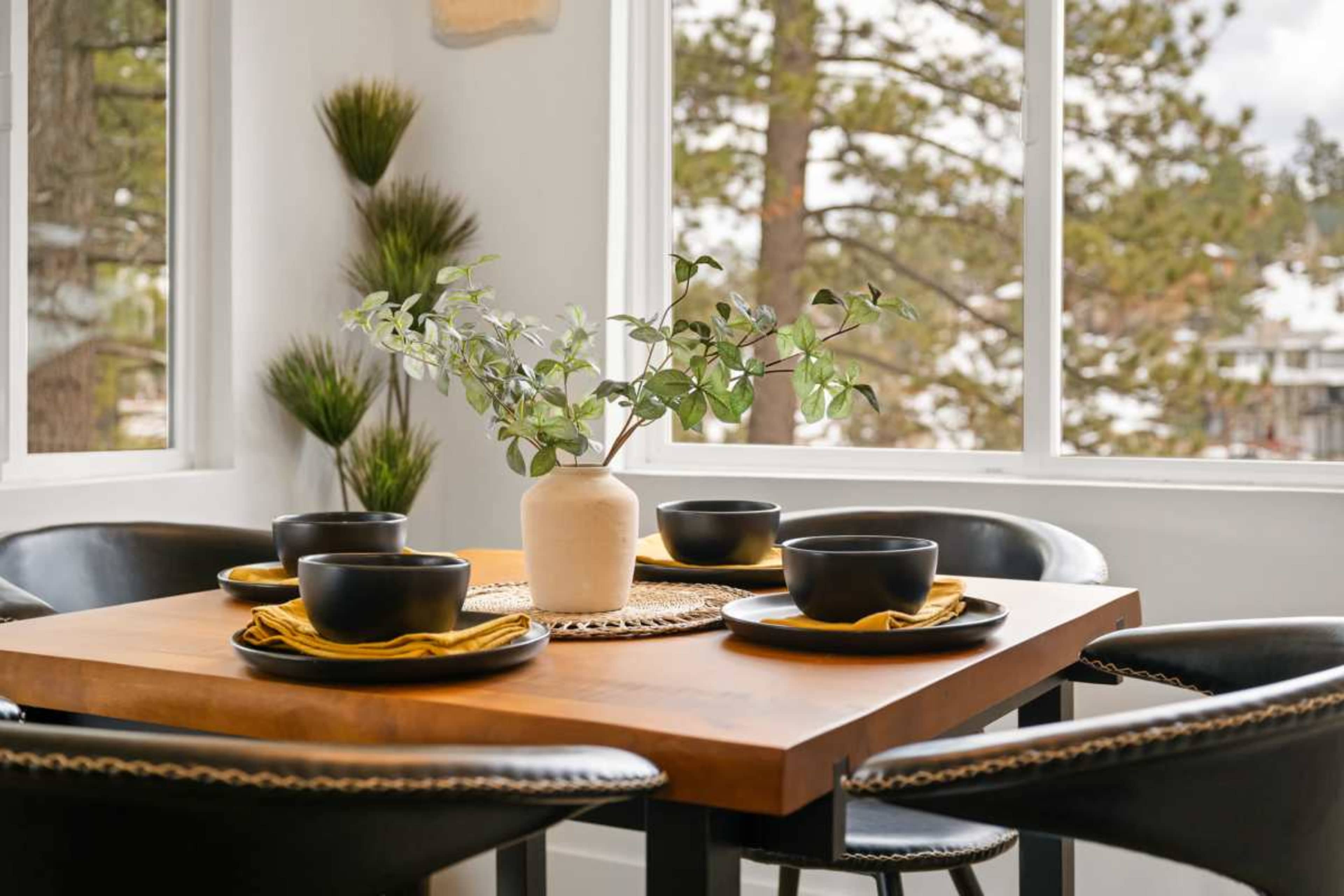 A wooden dining table set with black bowls and yellow napkins, surrounded by chairs, features a vase with greenery and large windows overlooking a treed landscape.