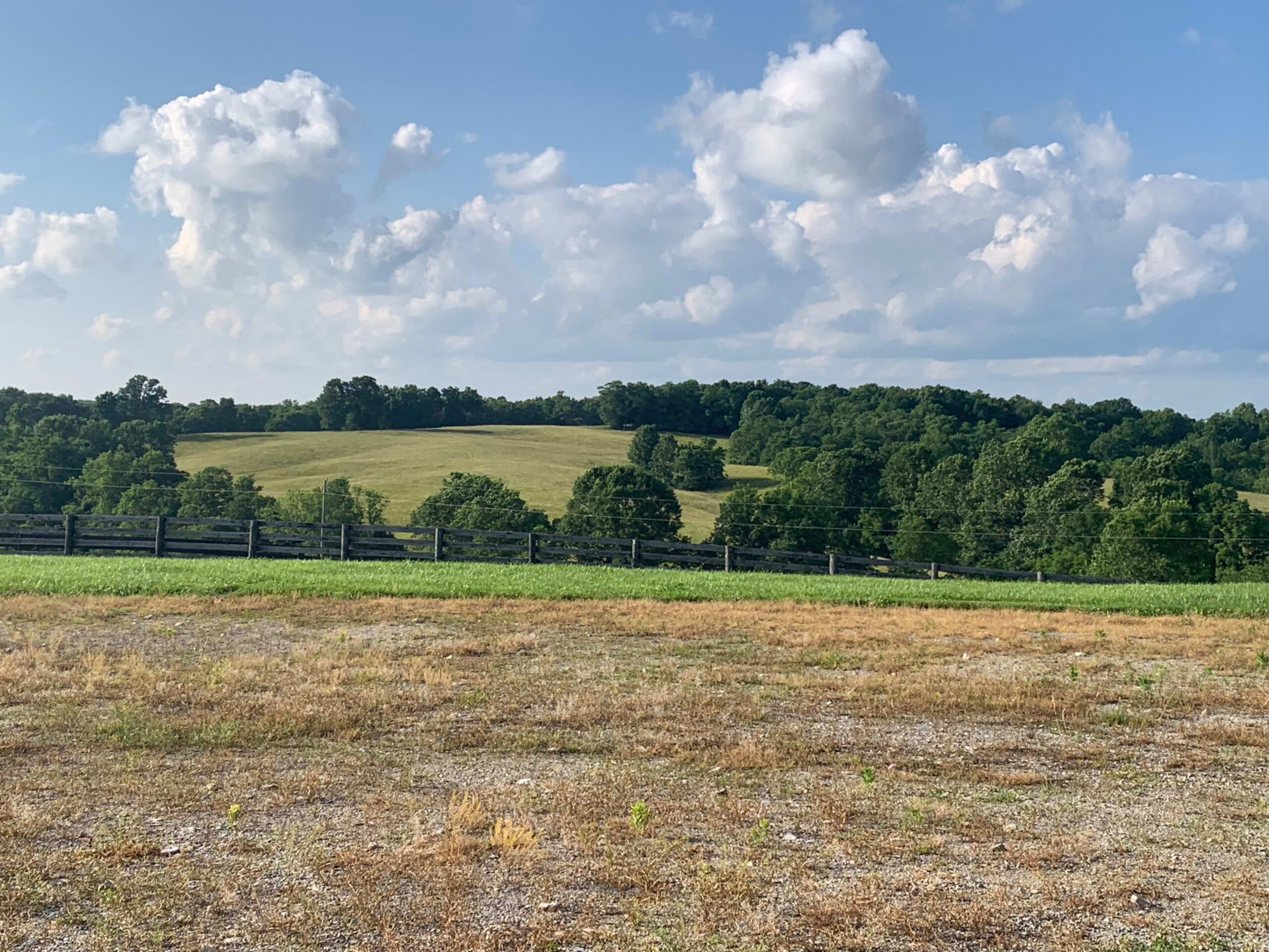 The image shows a grassy field with a wooden fence in the foreground, leading to rolling hills covered in trees under a partly cloudy sky.