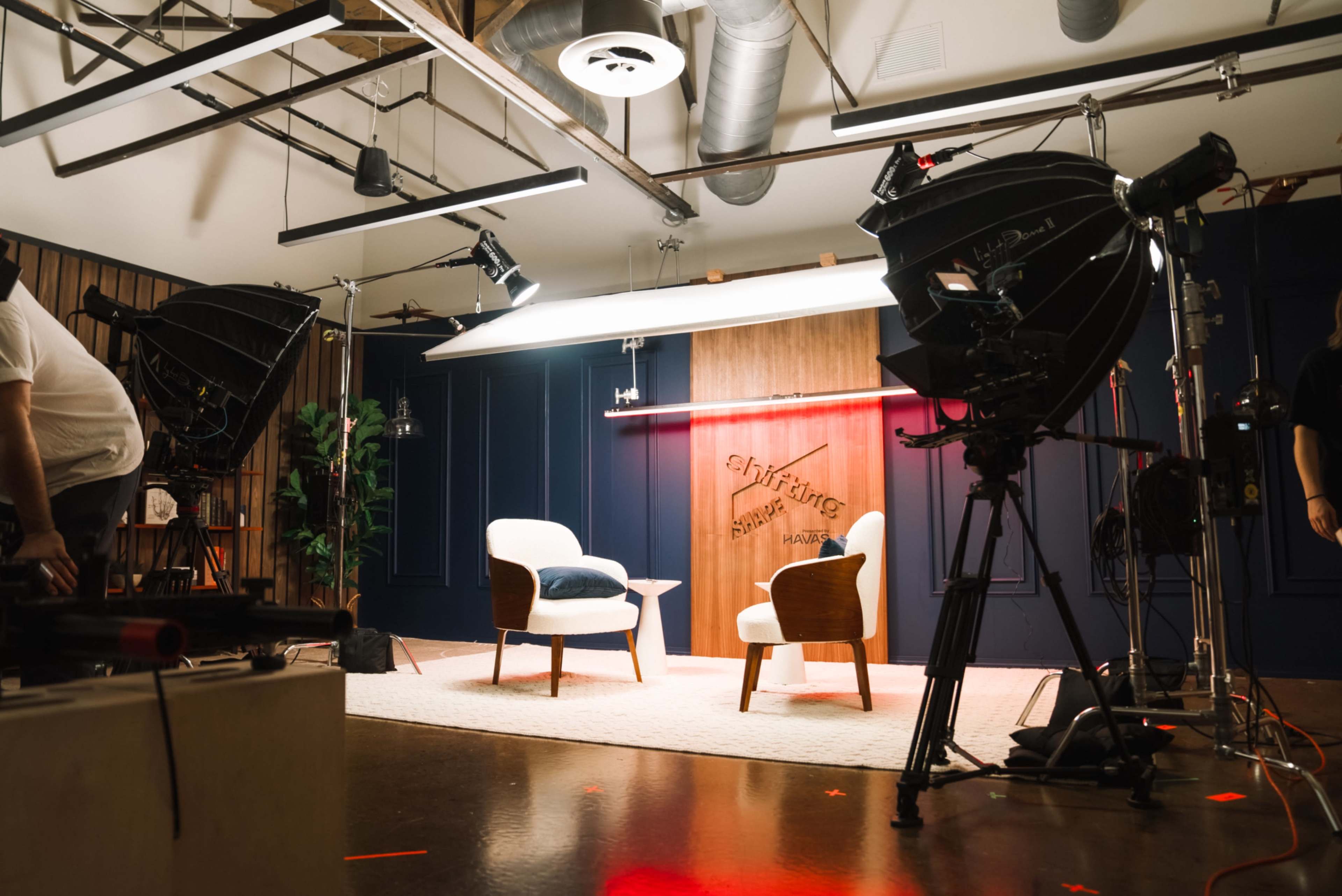 The image shows a television studio setup with two chairs on a carpet, surrounded by various lighting equipment and cameras.