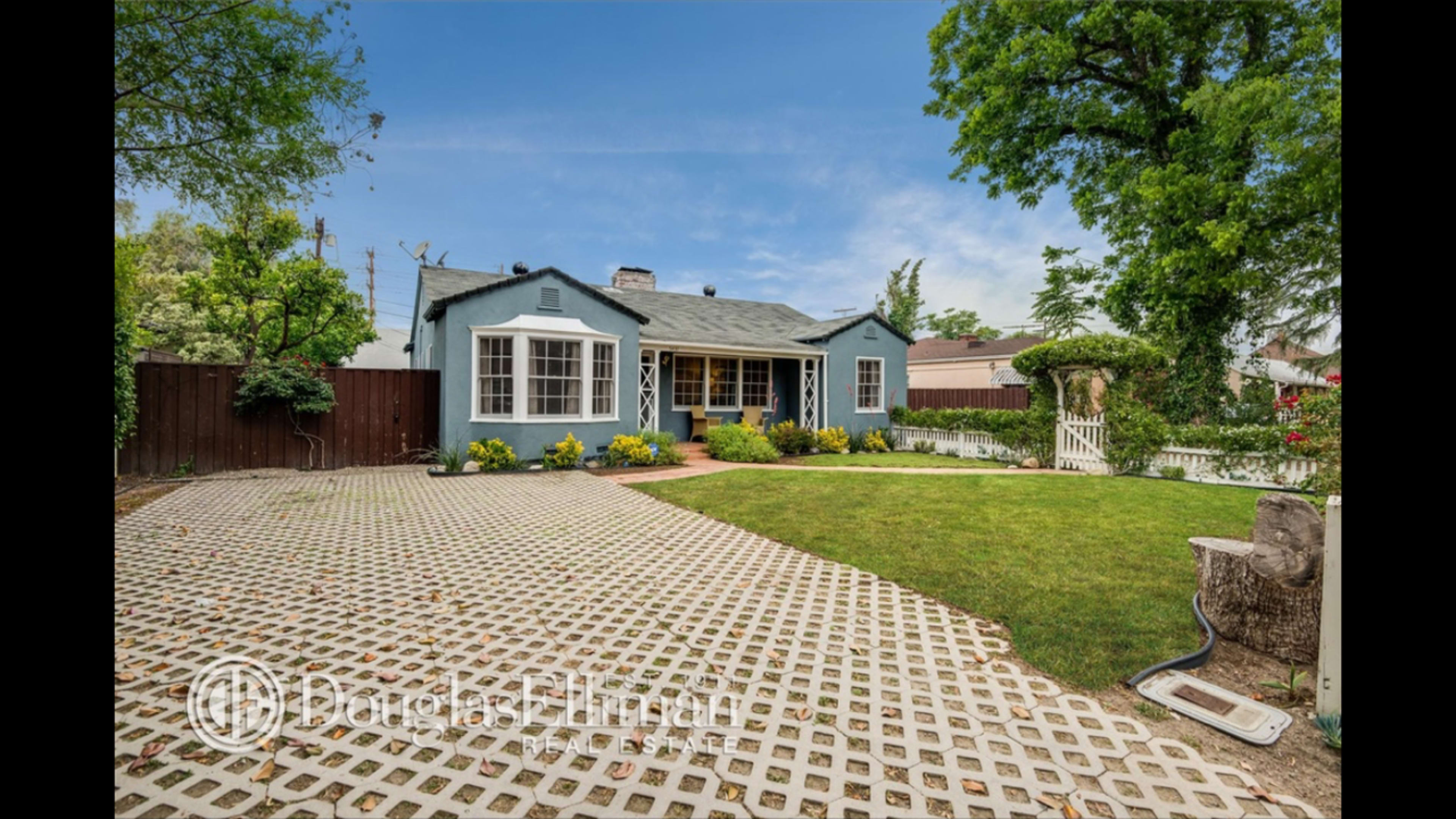 A light blue house with a front yard featuring a well-maintained lawn, surrounded by a white picket fence and flowering plants.
