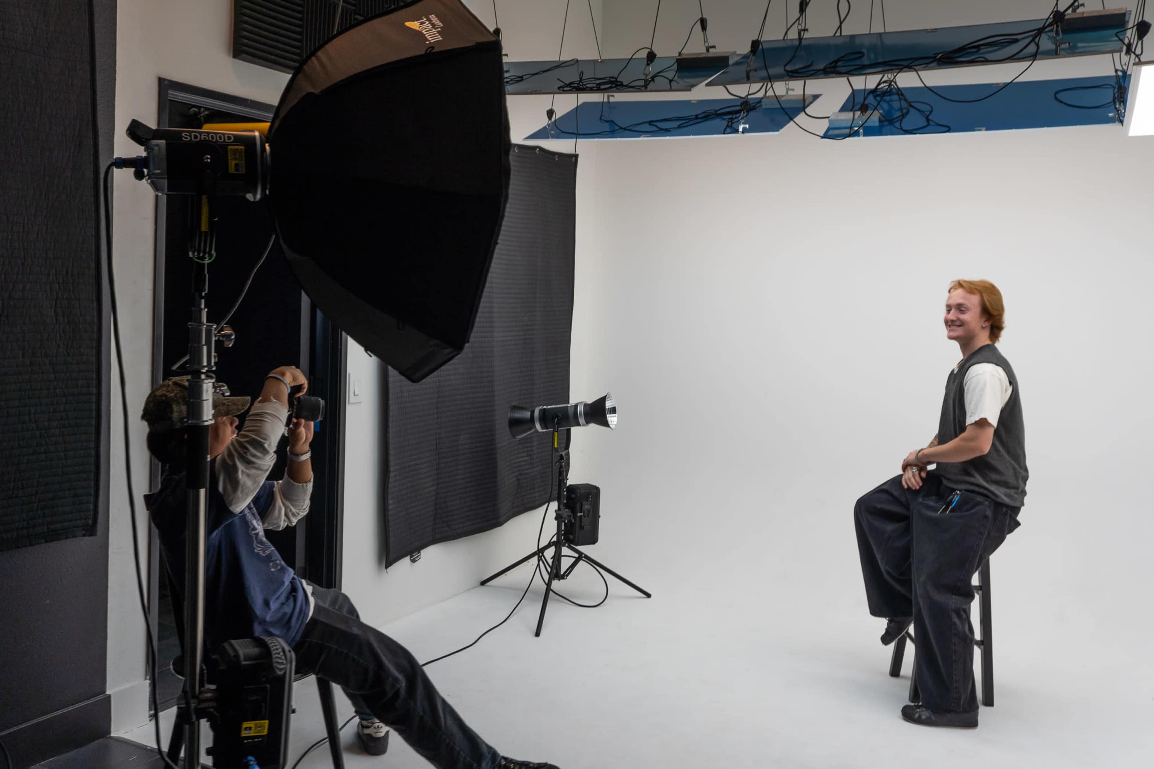 A photographer is setting up lighting equipment in a studio while a model poses on a stool against a plain white backdrop.