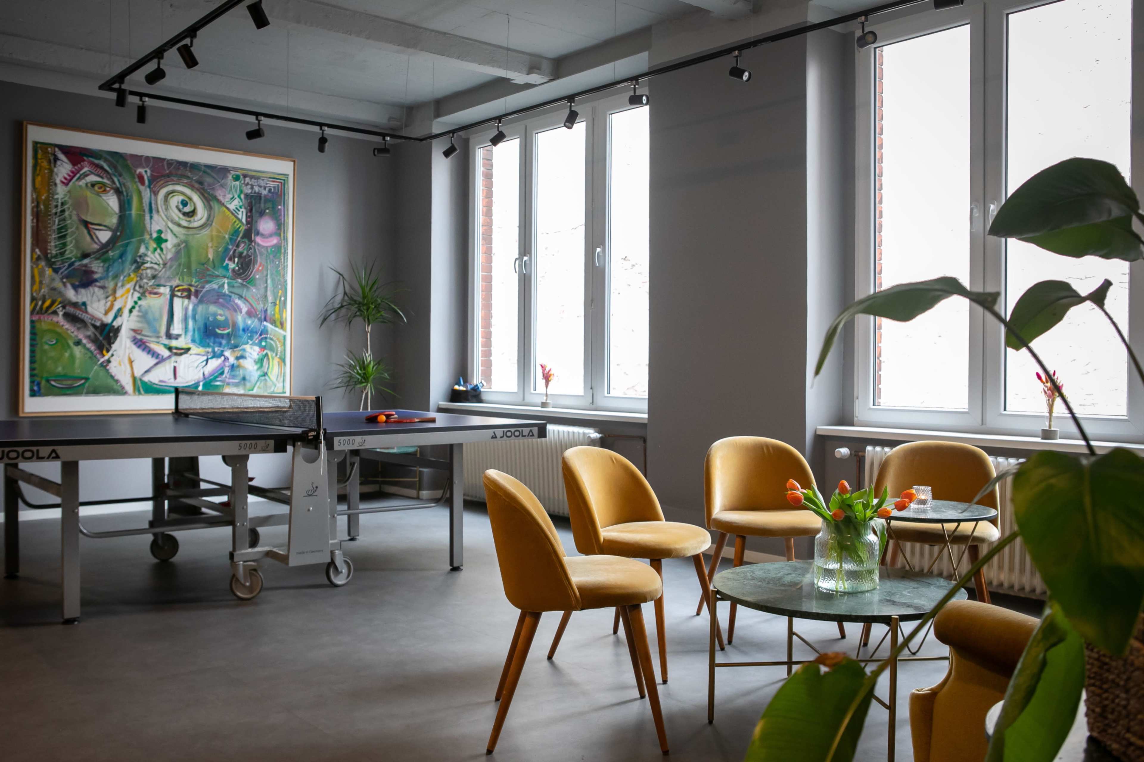 The image shows a modern room with a ping pong table, a colorful piece of artwork on the wall, and yellow chairs arranged around glass tables.