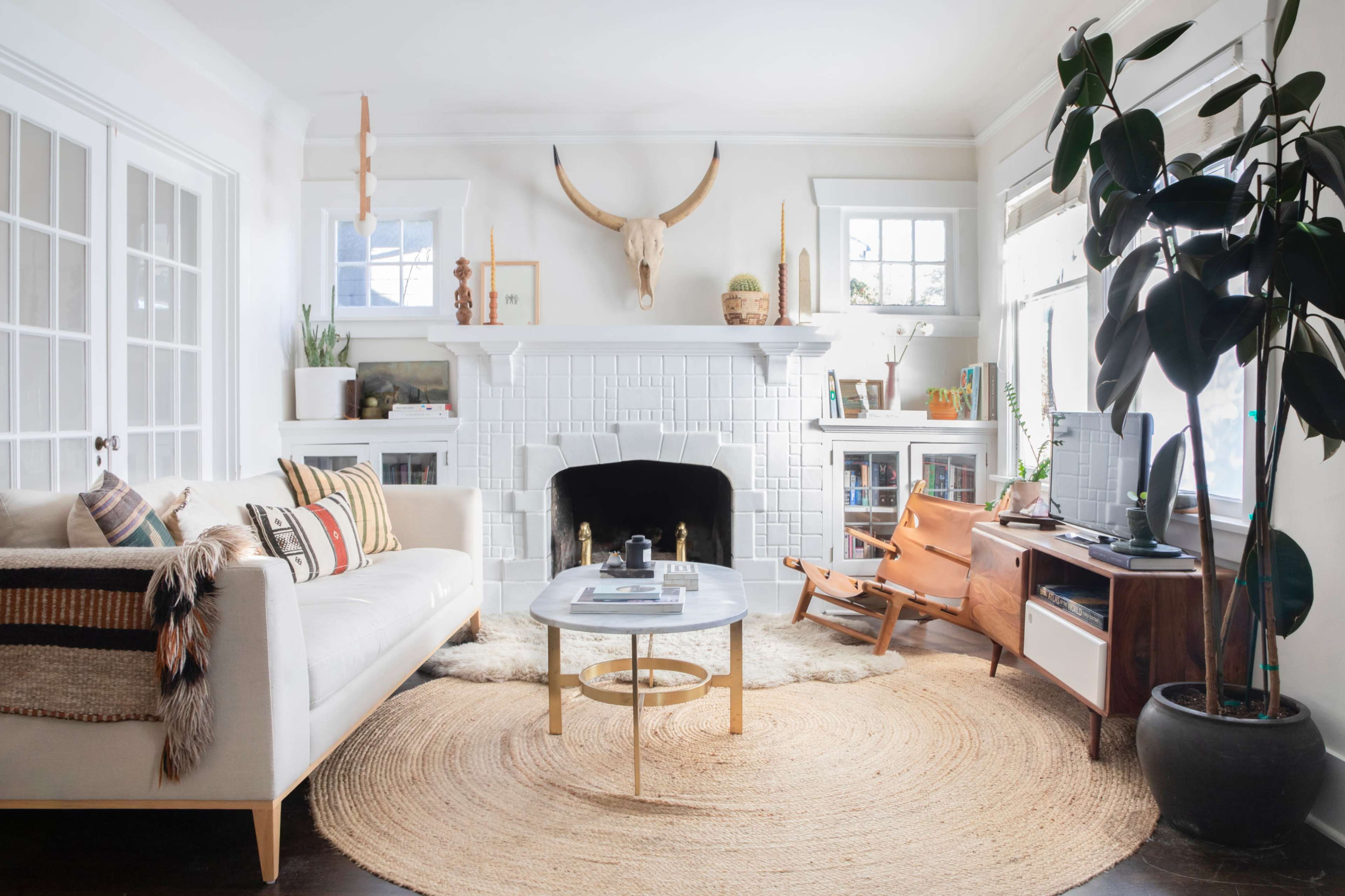 A bright living room features a white sofa, a round coffee table, a tan rug, and a fireplace adorned with decorative items and a mounted animal skull.