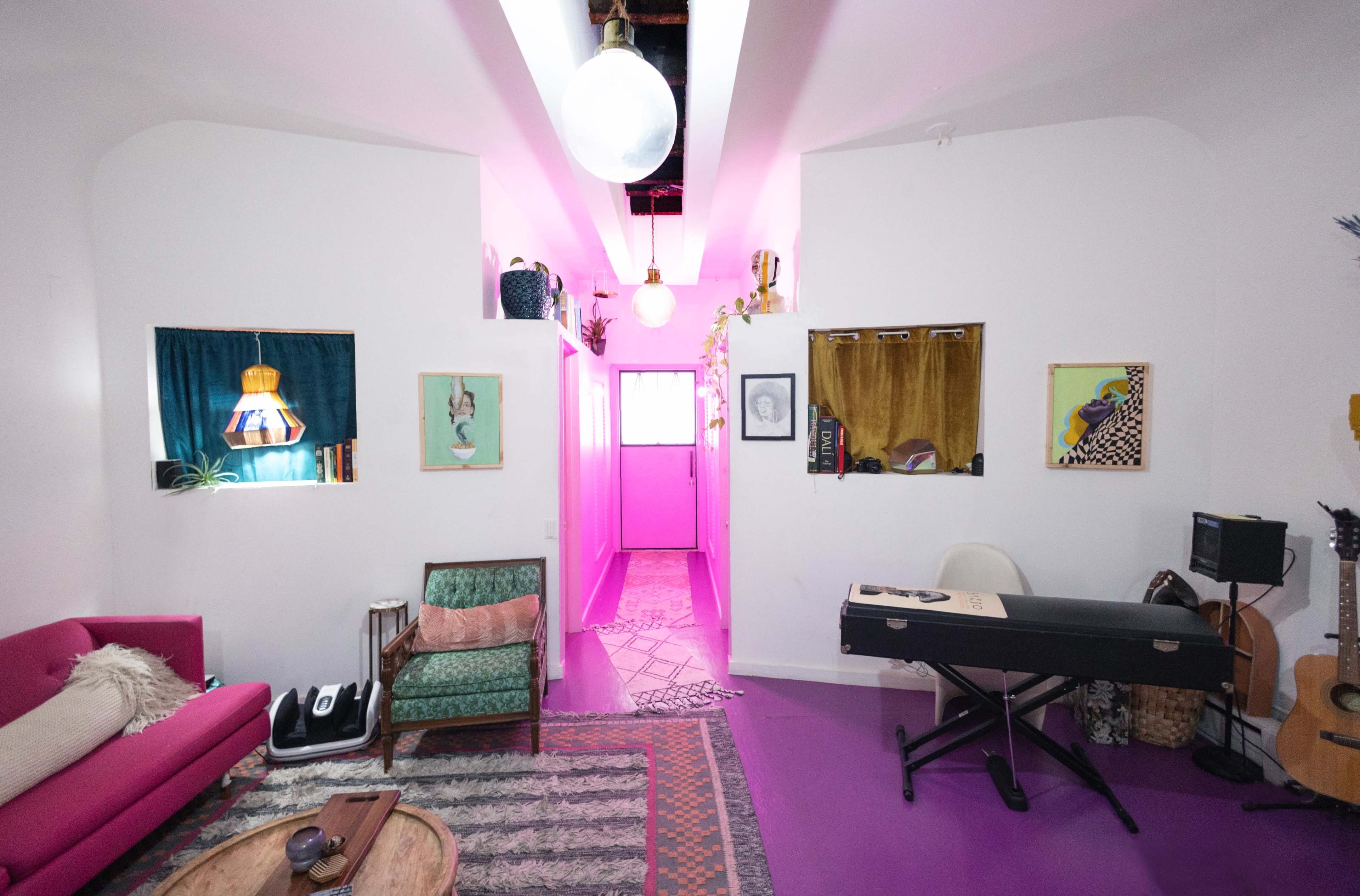 The image shows a brightly colored living space with a pink floor, a pink couch, and artistic decorations on the walls.