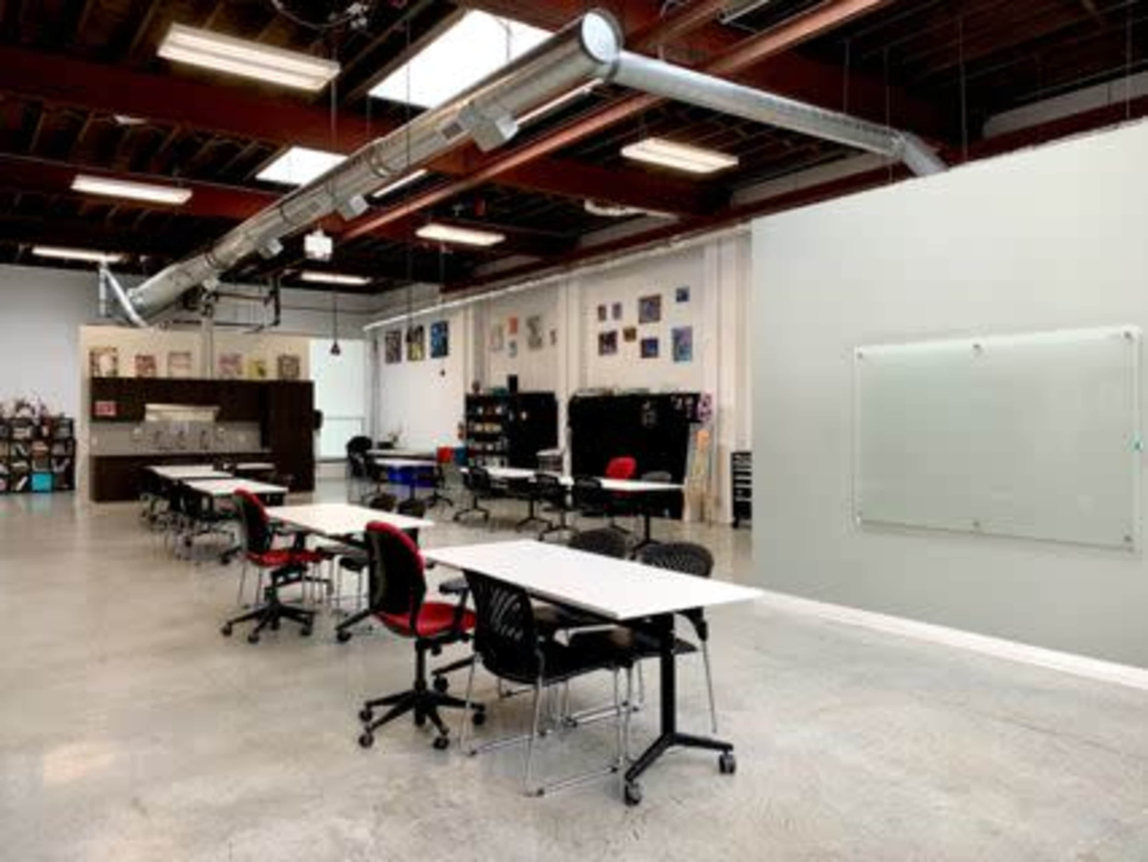 An open workspace features several tables and chairs, along with a large whiteboard and exposed ductwork on the ceiling.