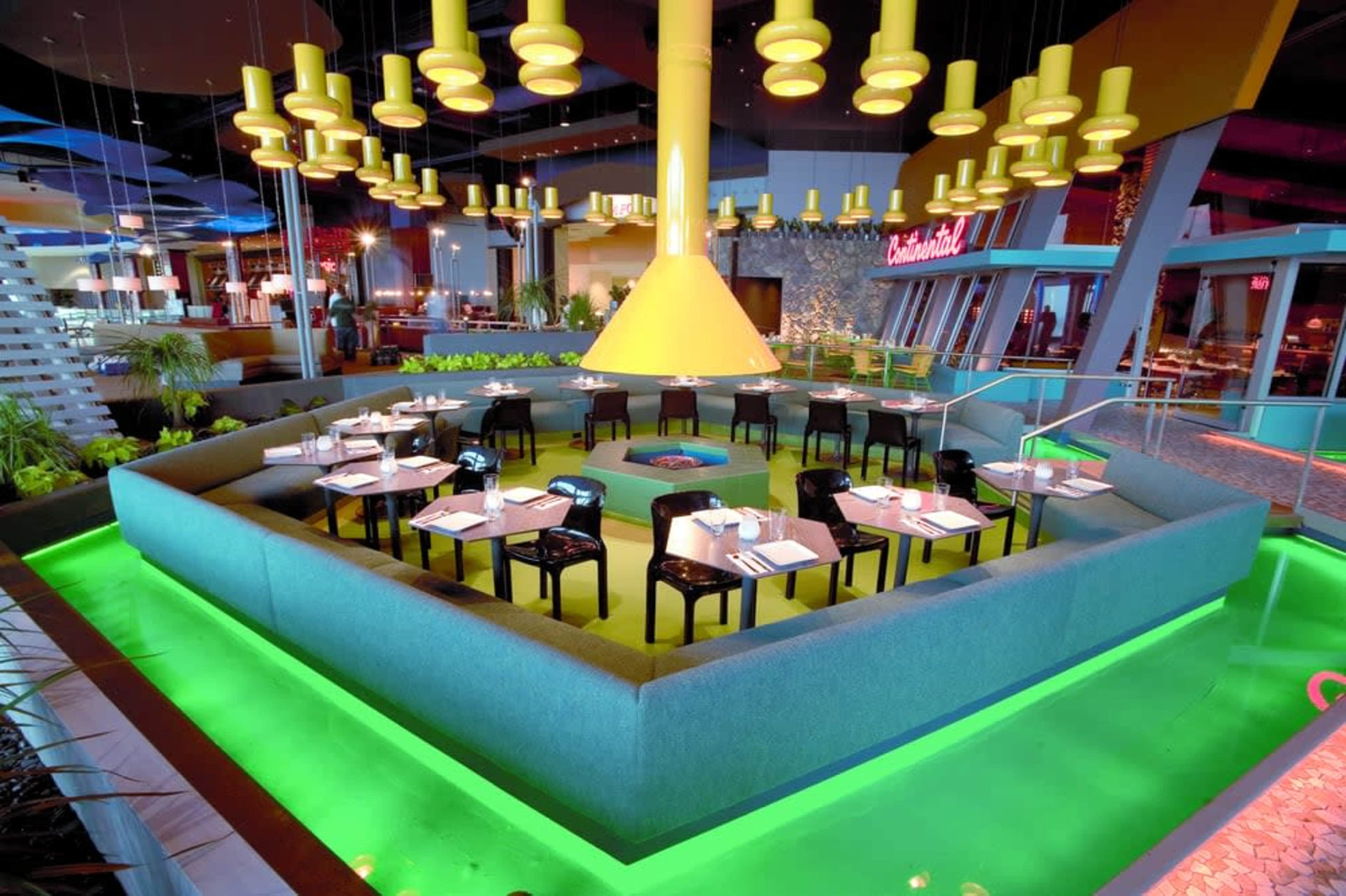 A modern restaurant interior with a large, central table surrounded by seating and illuminated by pendant lights.