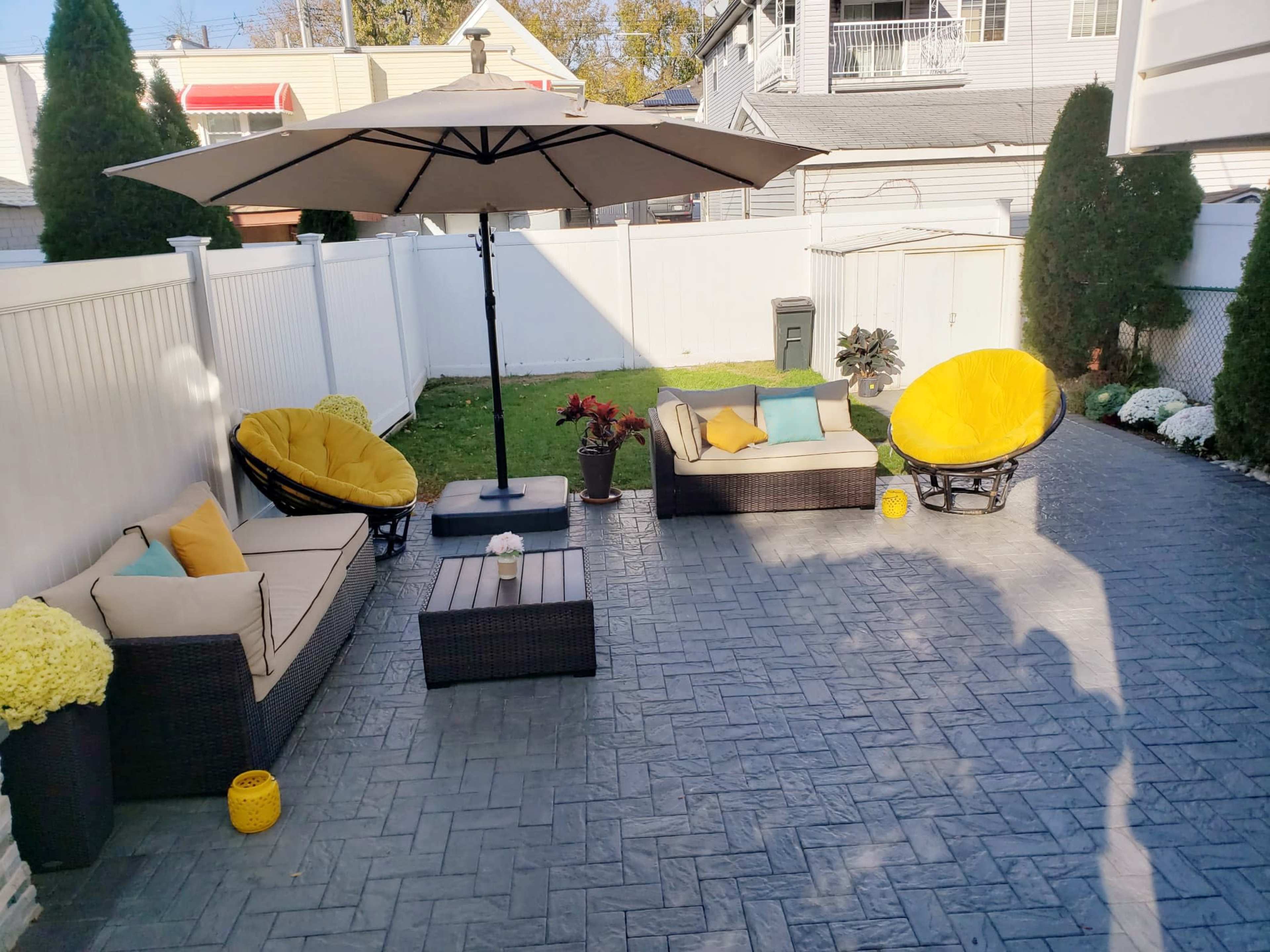 The image shows a backyard patio with two yellow cushioned lounge chairs, two beige sofas, a central coffee table, and a large umbrella, surrounded by a white fence and greenery.