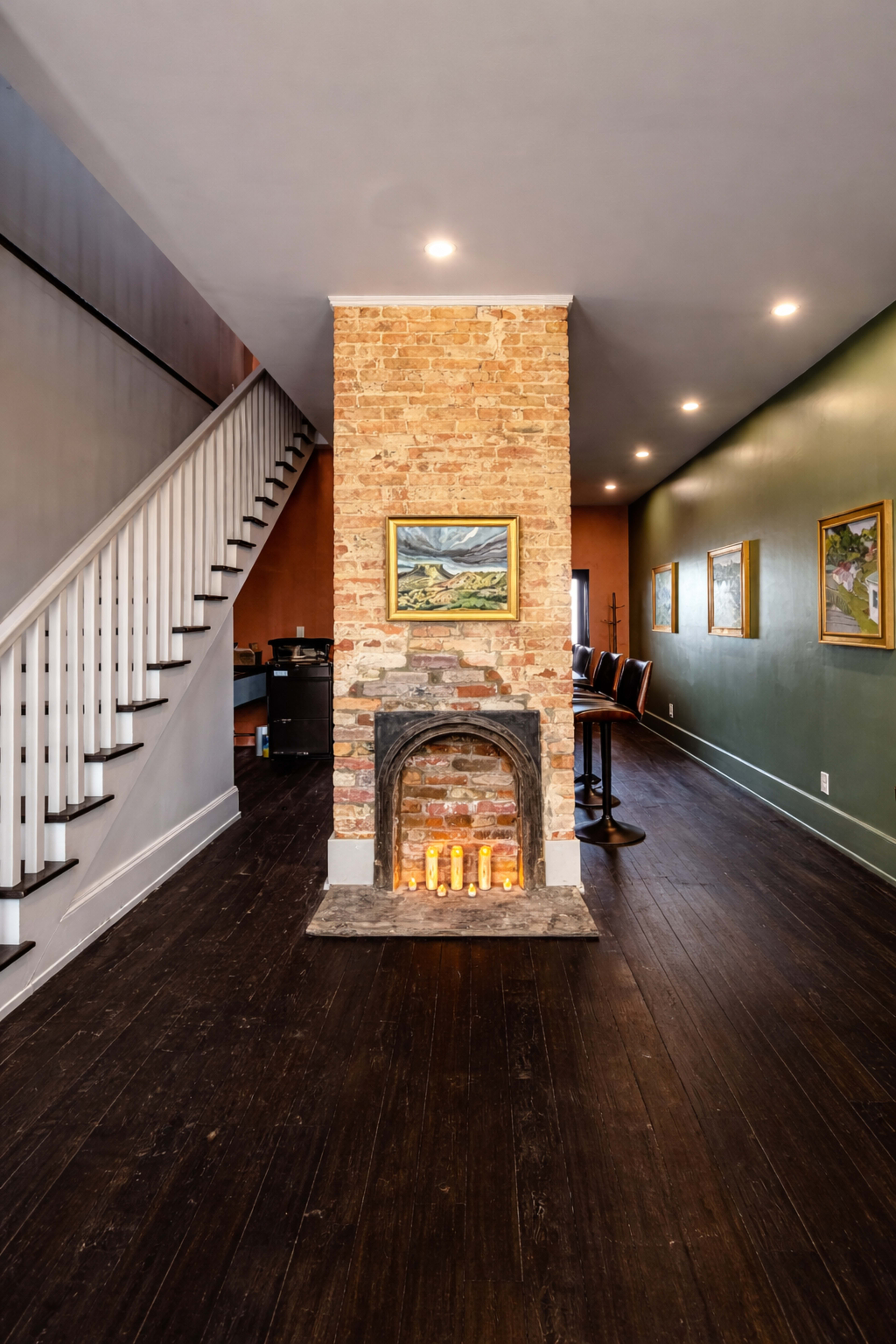 The image shows an interior space featuring a brick fireplace at the center, with a staircase on the left and artwork hanging on a green wall.
