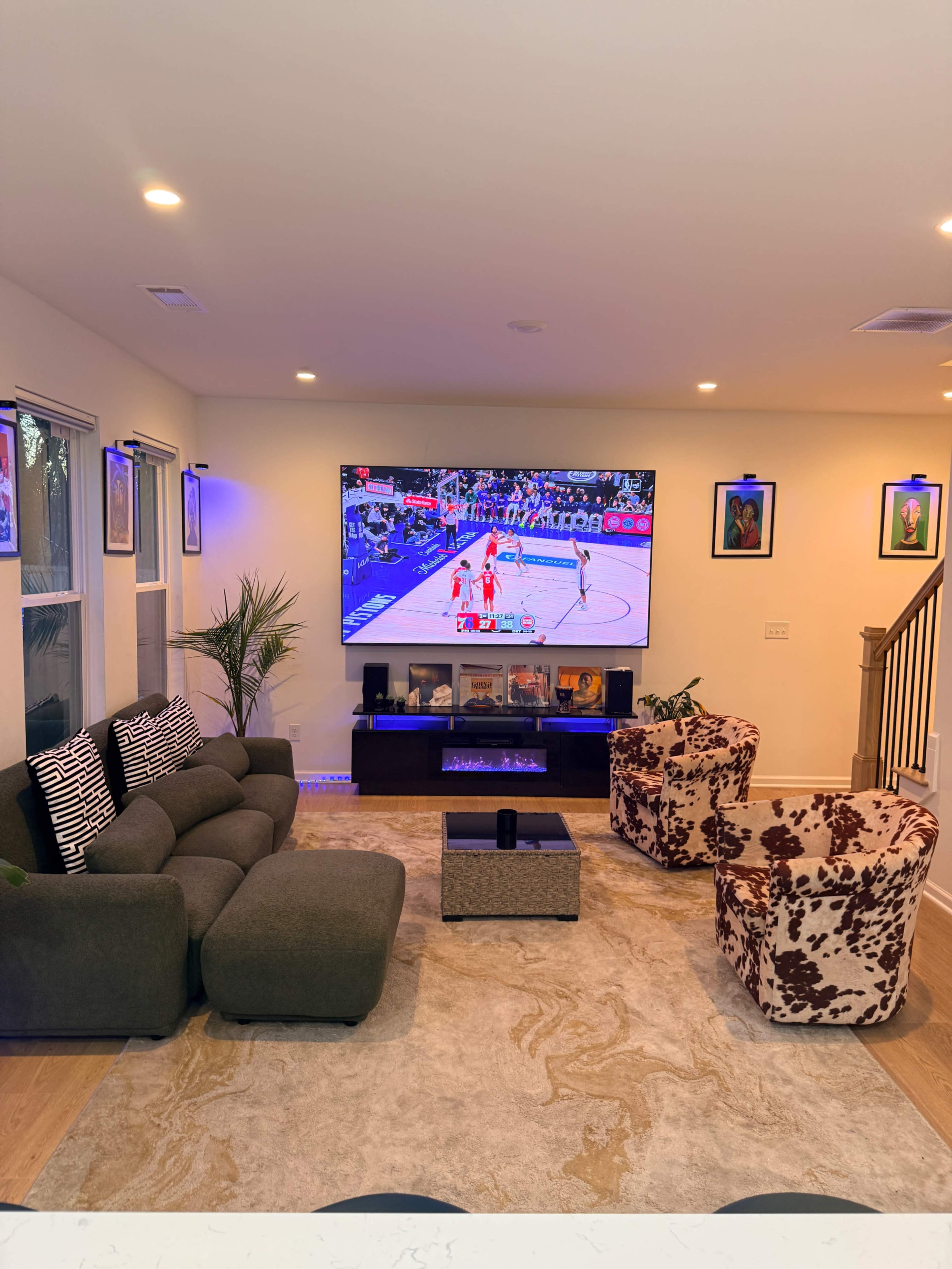 A living room features a large television displaying a basketball game, surrounded by comfortable seating and decorative plants.