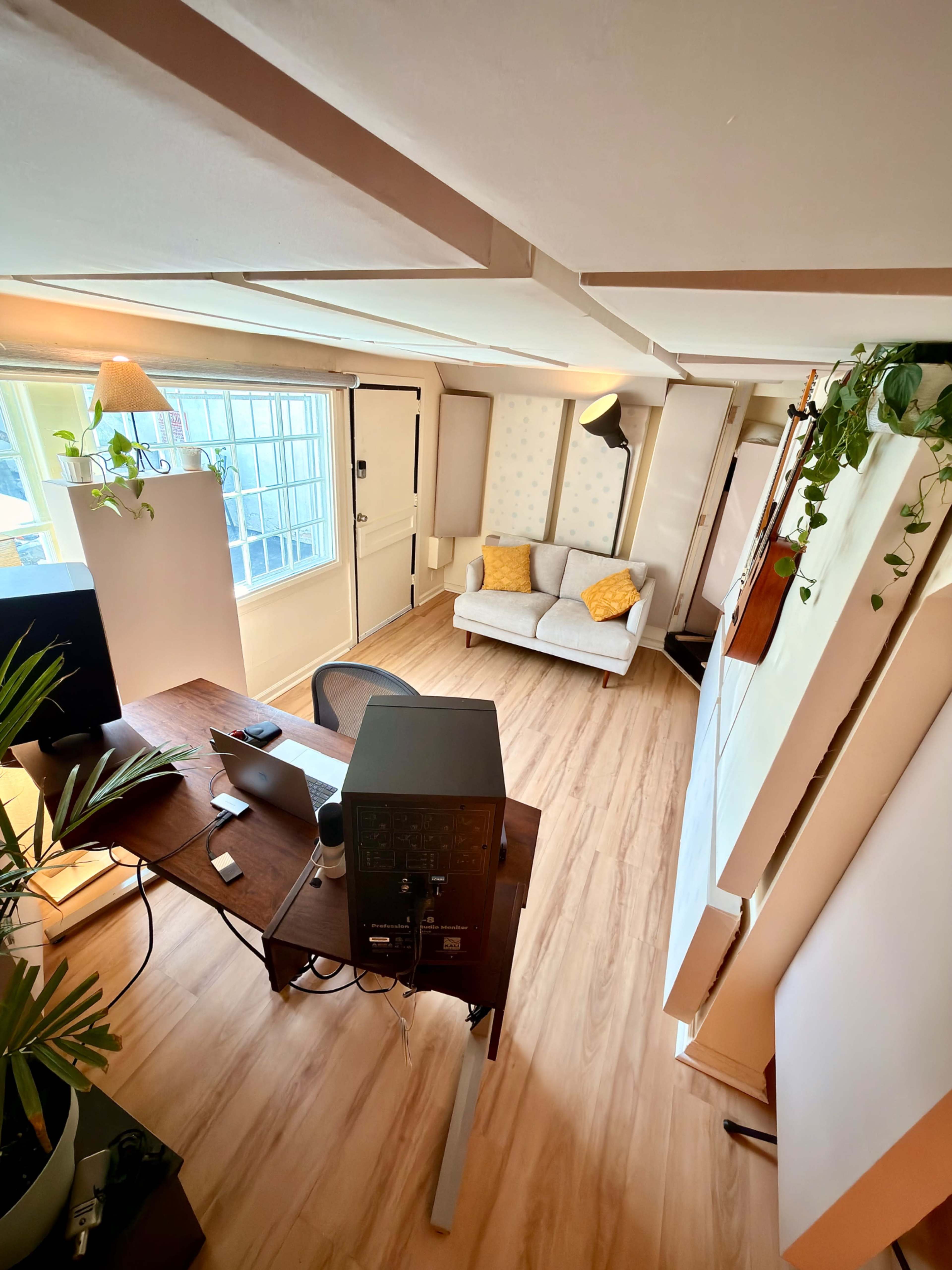 The image shows a bright office space featuring a desk with a computer, a gray sofa with yellow cushions, and large windows allowing natural light to enter.