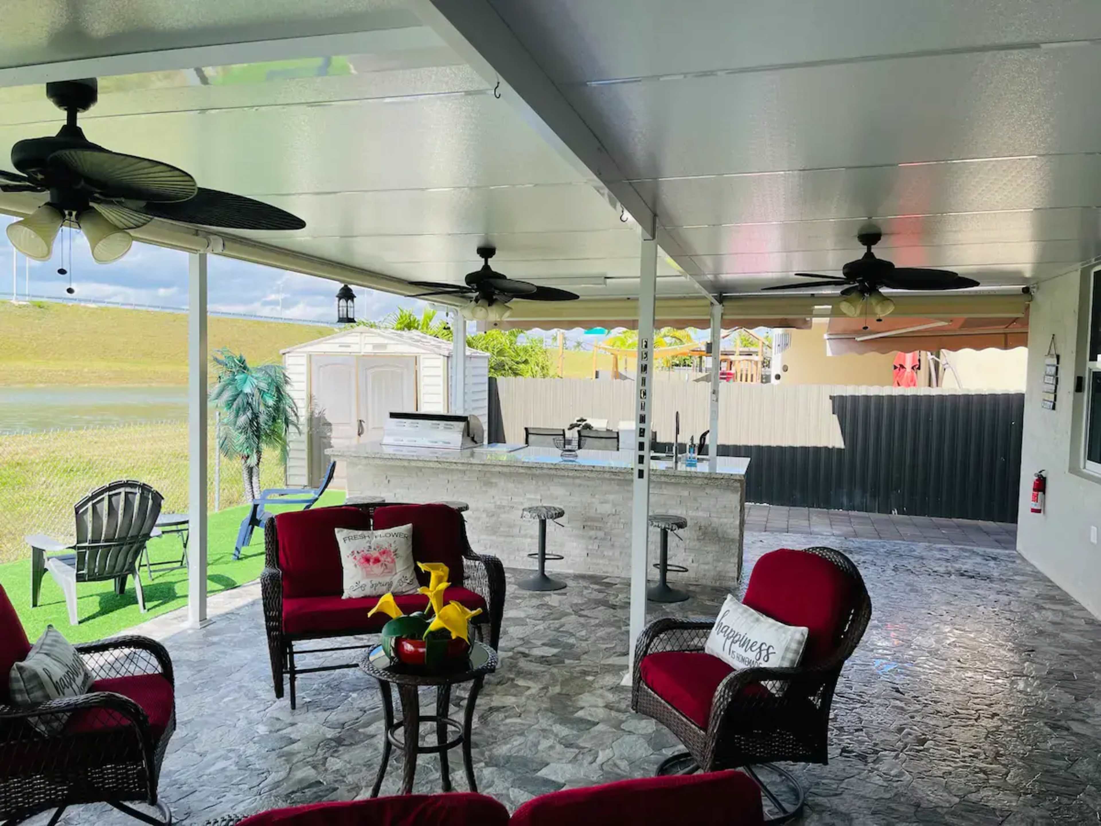 A covered outdoor patio with red seating, a small table, a bar area, and a well-maintained lawn.