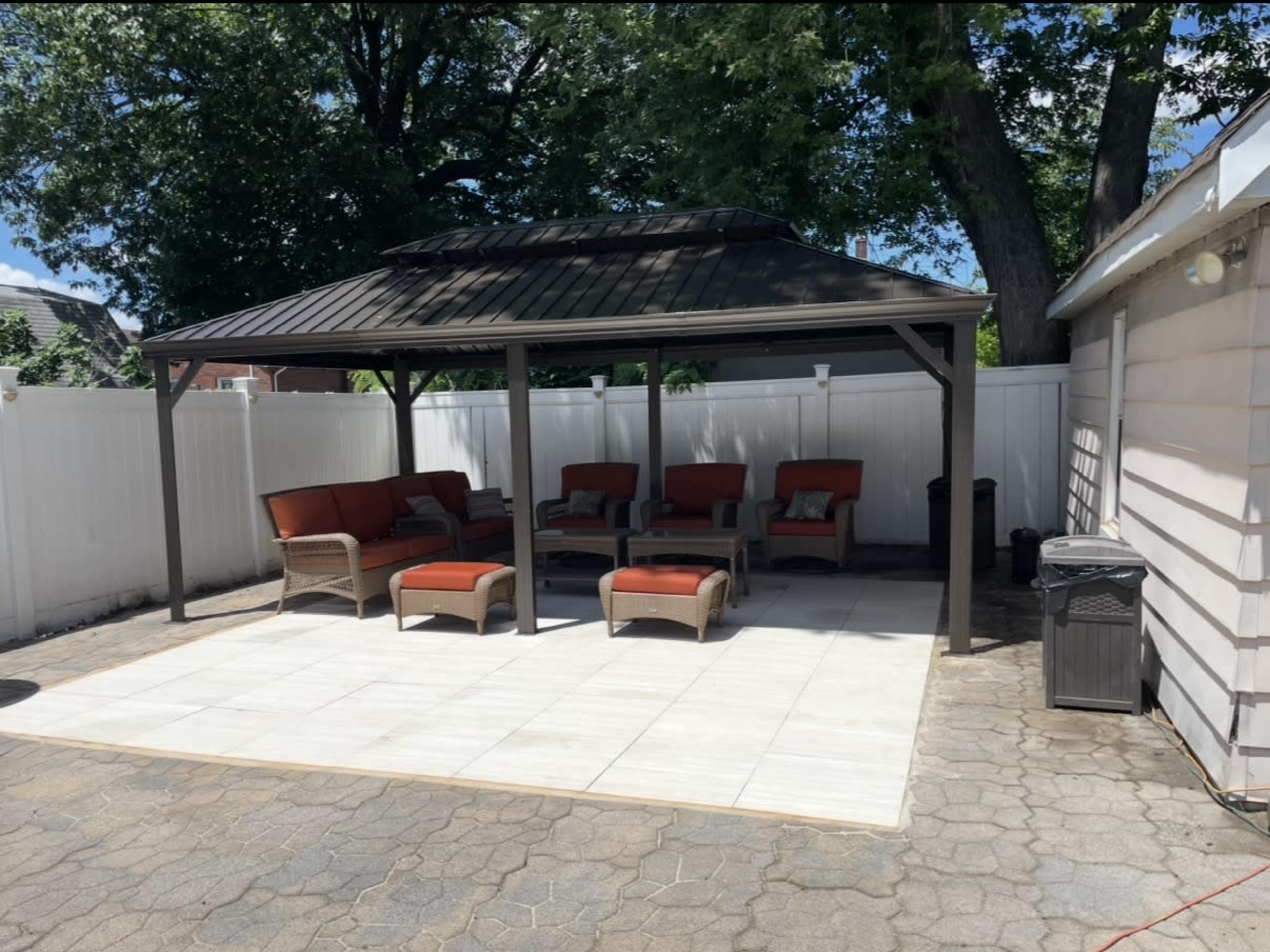 A covered outdoor seating area with a metal roof, featuring wicker furniture with orange cushions, is situated on a stone patio surrounded by a white fence and trees.