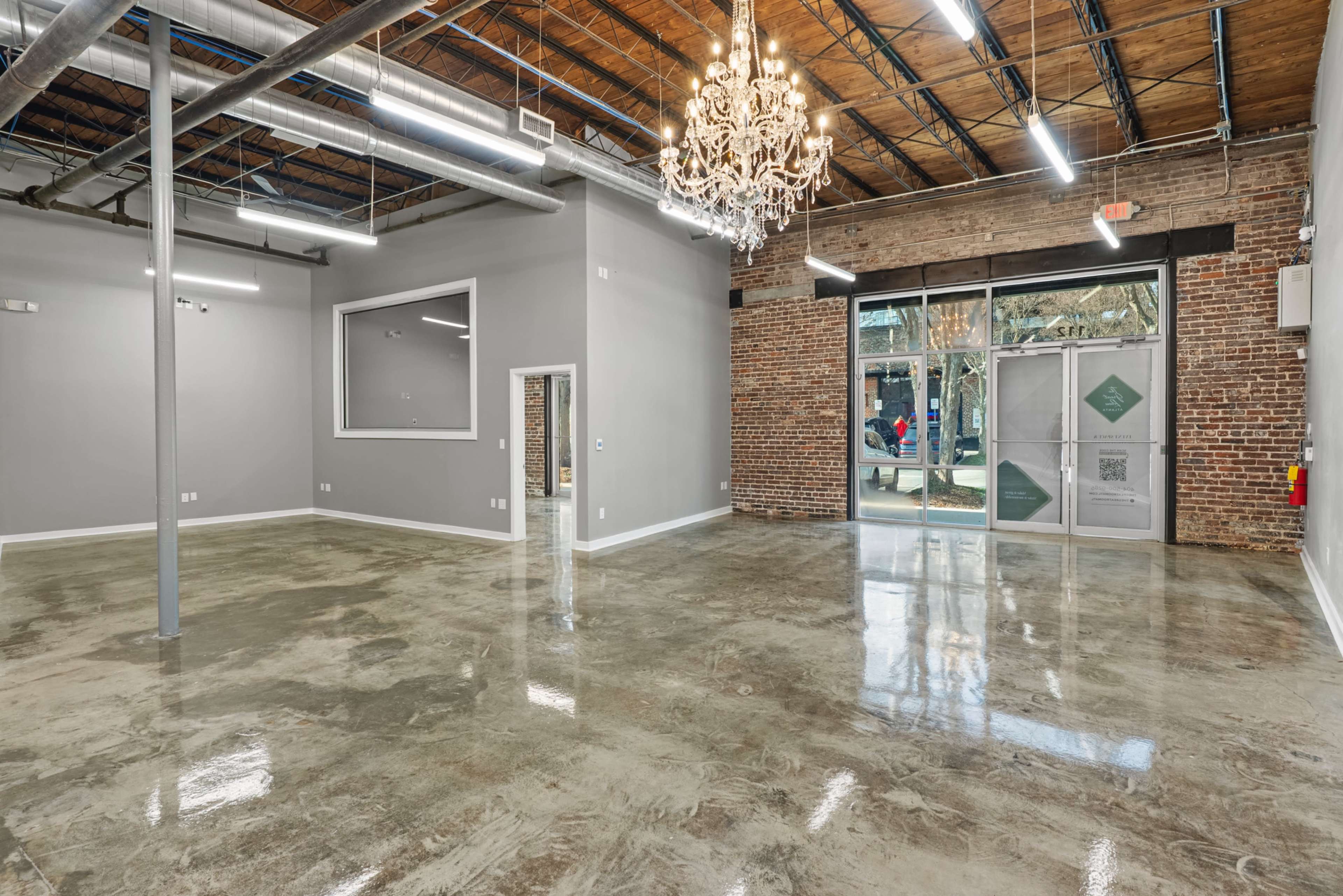 The image shows an empty commercial interior space with polished concrete floors, exposed brick walls, and a large glass entrance featuring a chandelier above.