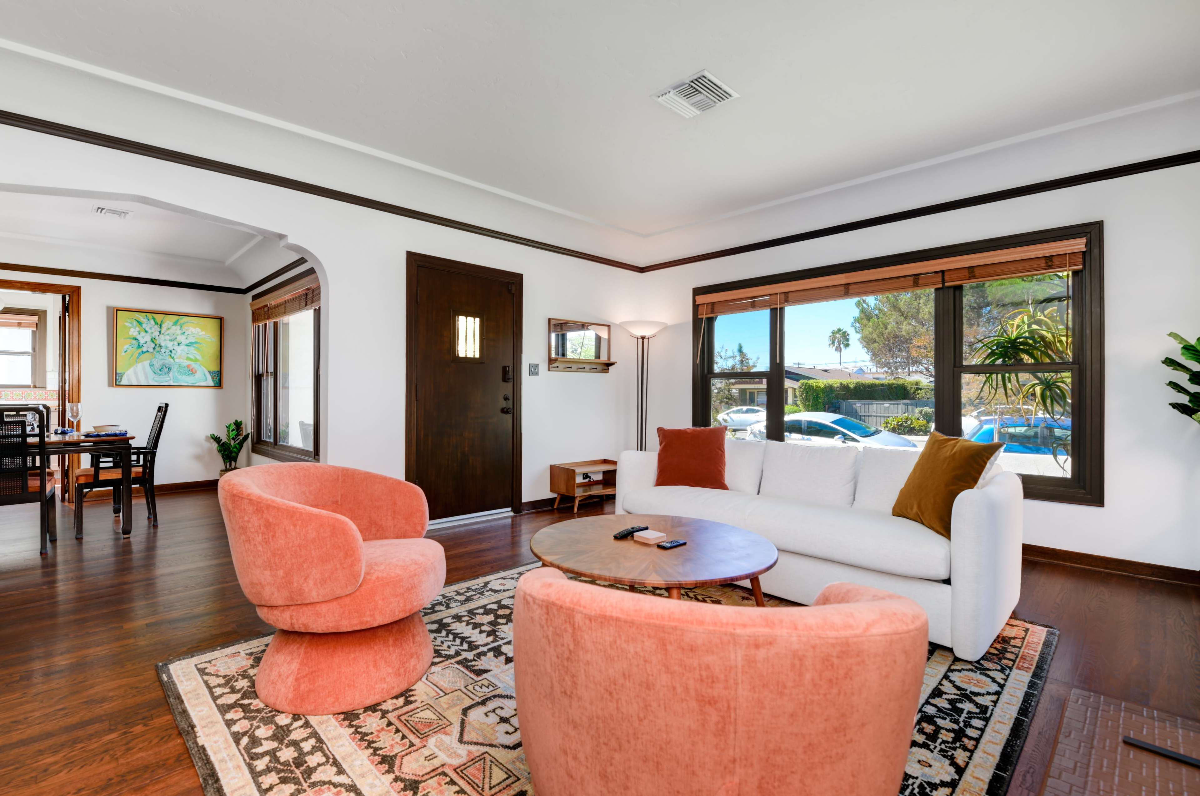 The cozy living room features two pink armchairs and a white sofa, with a round coffee table in the center and large windows letting in natural light.