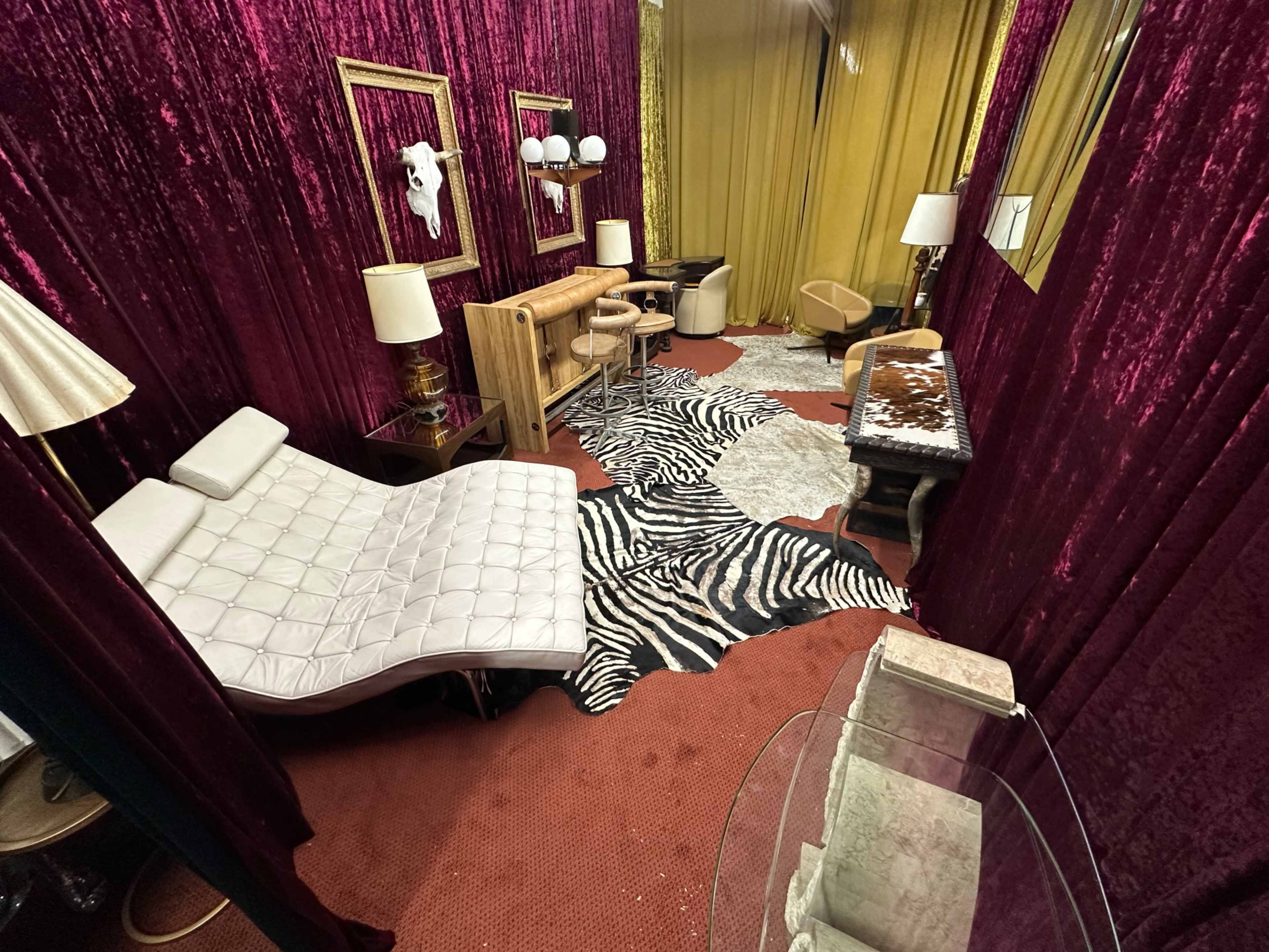 The image depicts a small room with red velvet walls, featuring a white chaise lounge, zebra-striped rug, and various lamps, alongside a gold curtain backdrop.