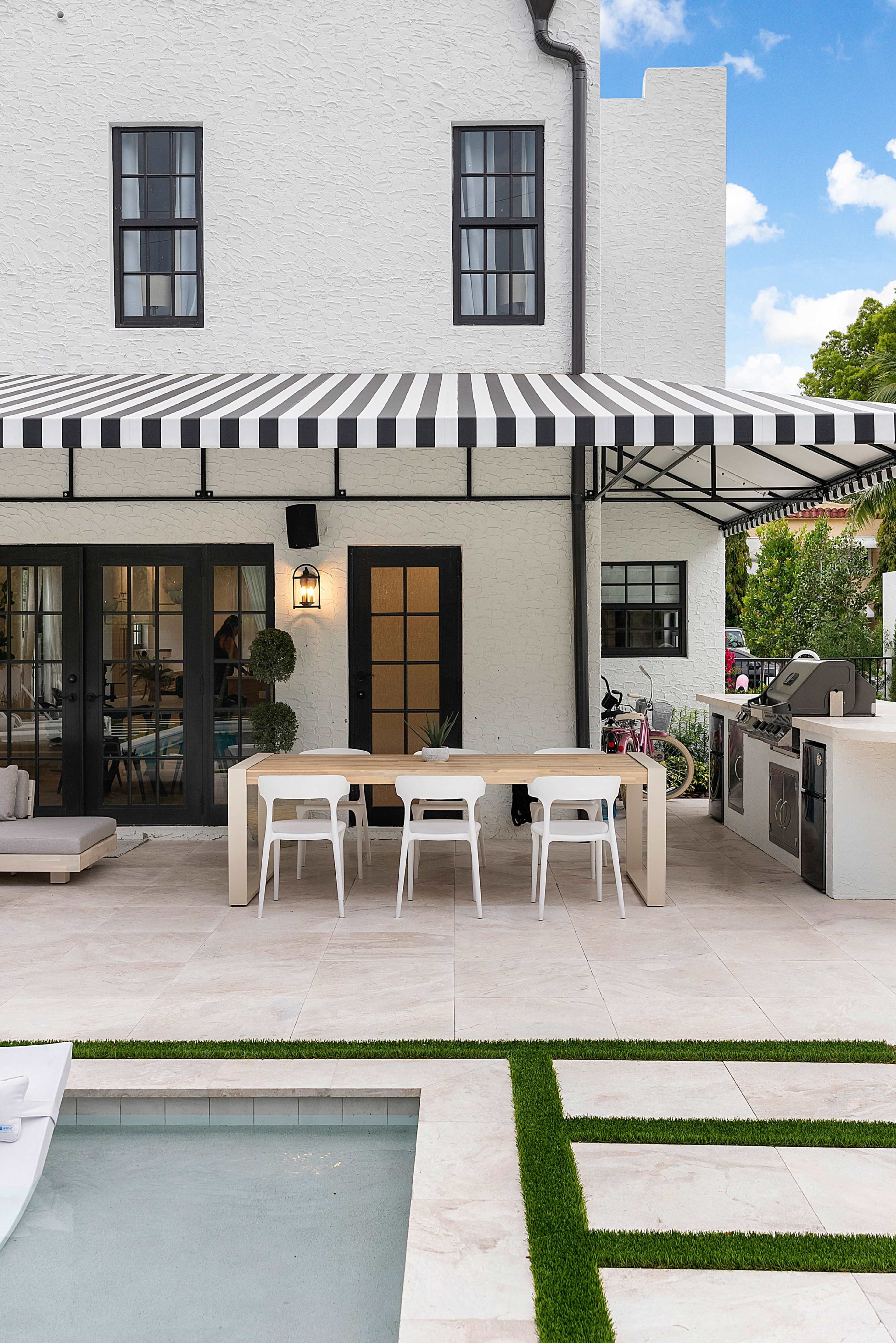 The image shows a modern outdoor dining area with a striped awning, a rectangular table, and white chairs beside a swimming pool and landscaped grass.
