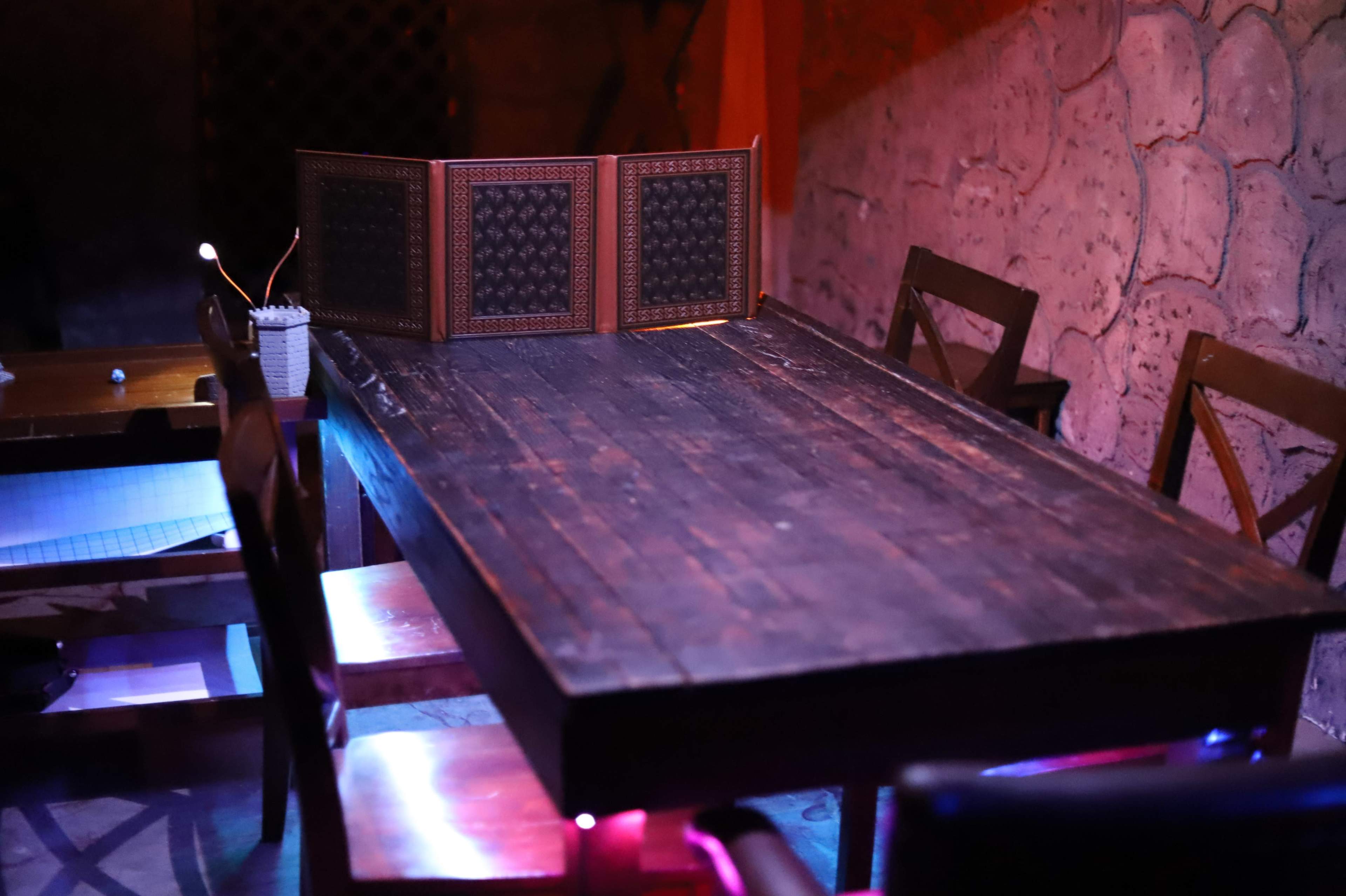 A large wooden table with a decorative folding screen sits in a dimly lit room with stone walls and surrounding chairs.