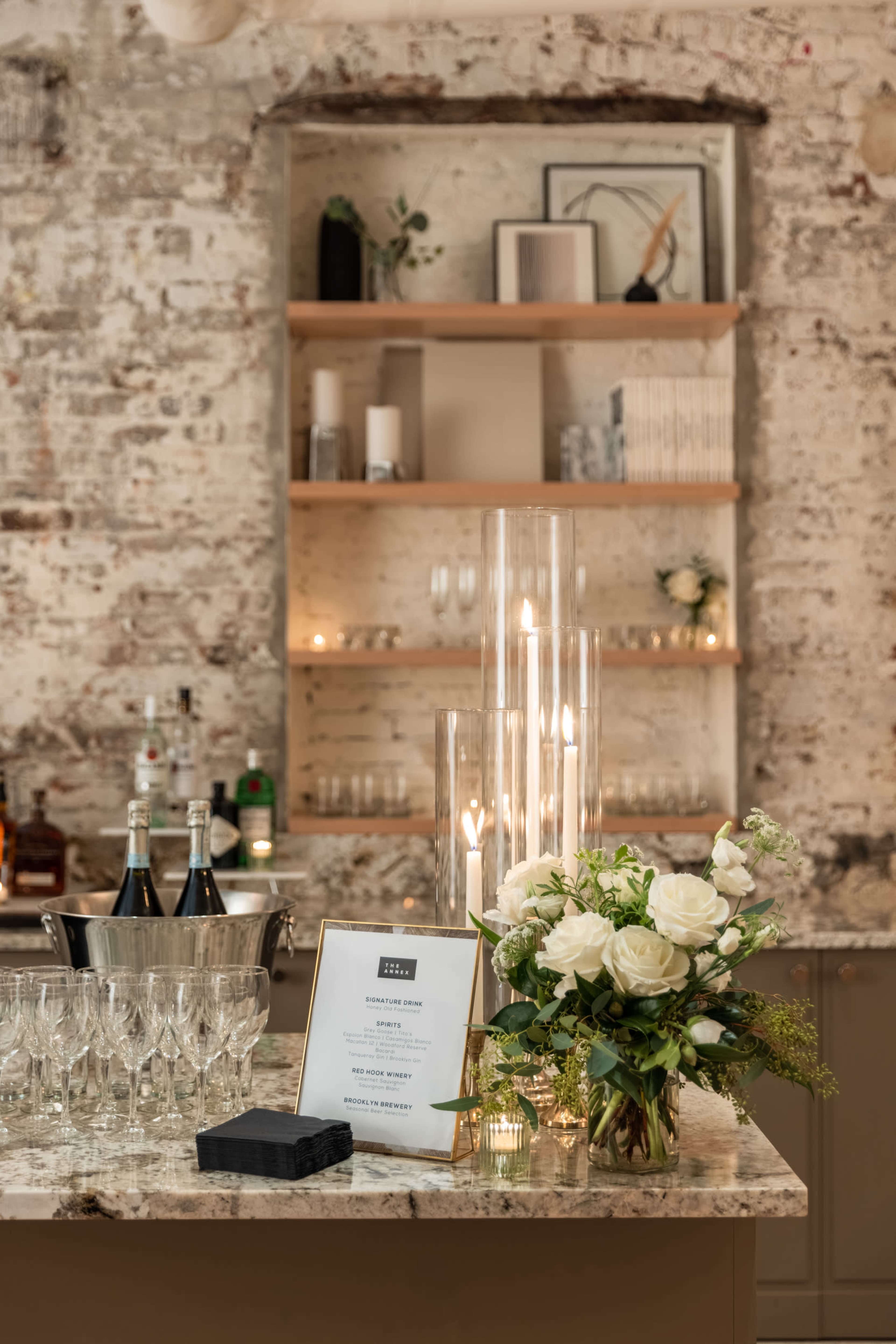 A bar area features a marble countertop with champagne bottles, glasses, a floral arrangement, and a menu displayed on a stand, against a backdrop of a rustic brick wall and wooden shelves.