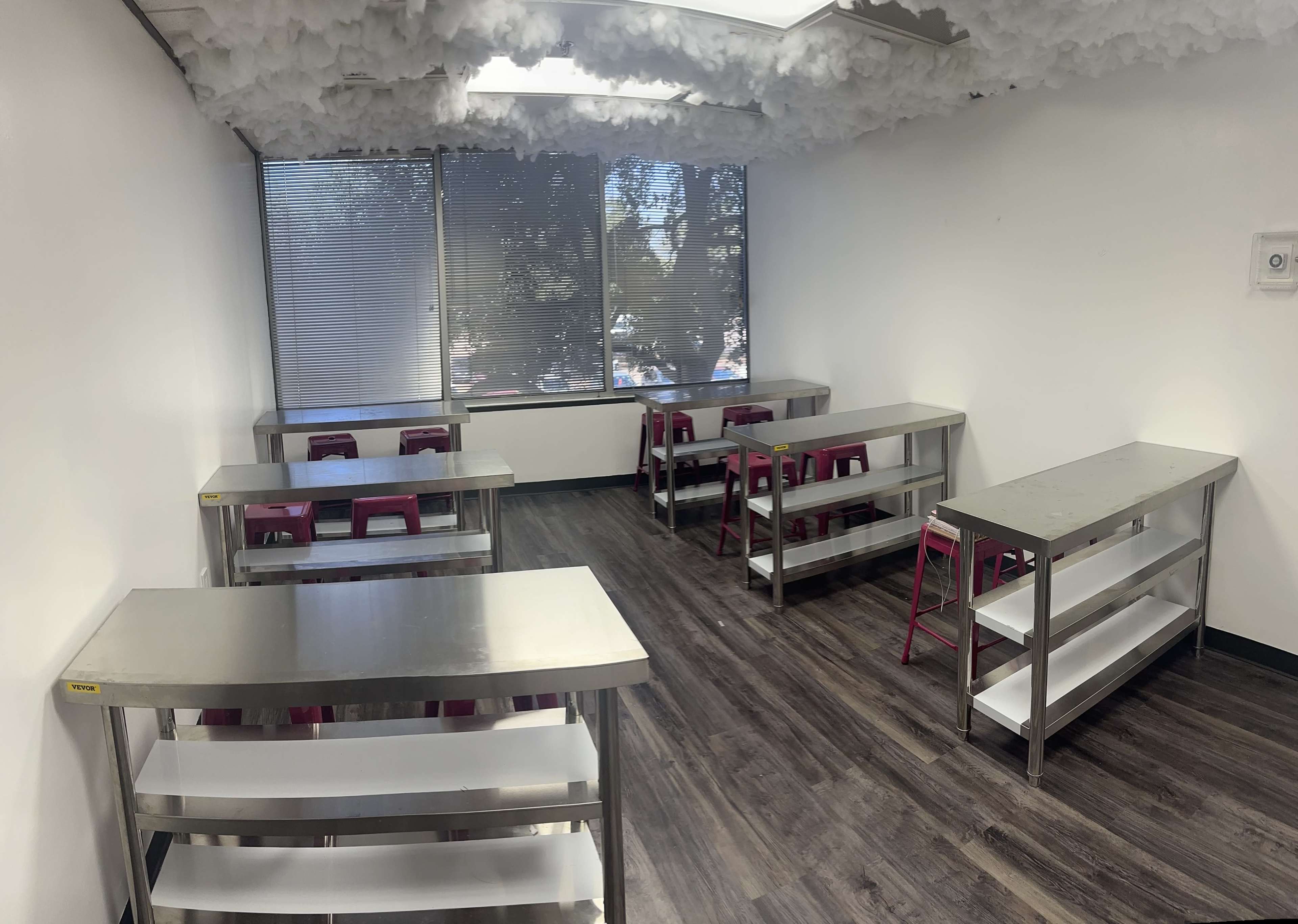 A classroom with multiple metal tables and pink chairs, featuring a ceiling decorated with cloud-like material and large windows.