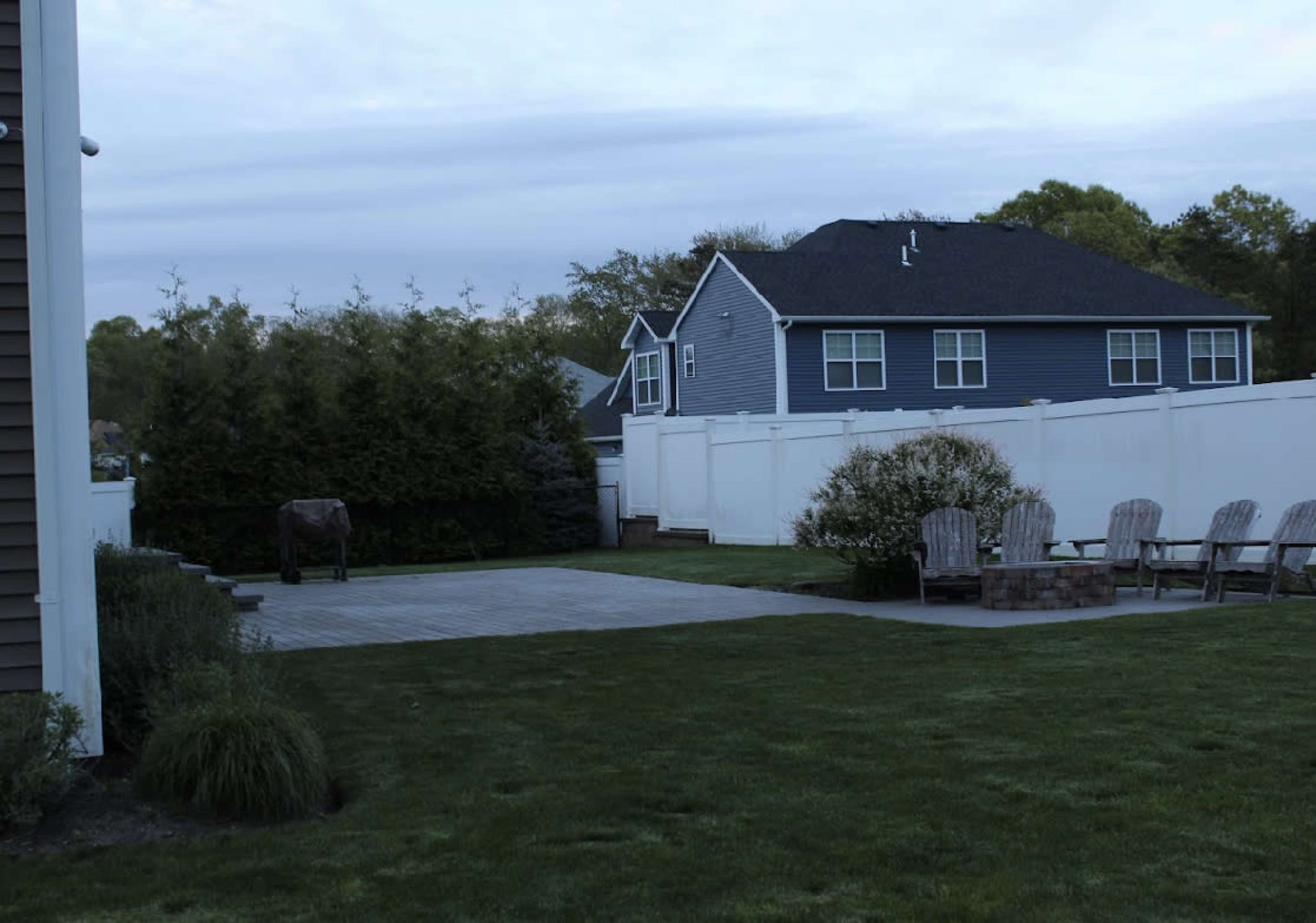 A clear backyard features a wooden patio, a grassy area, and several Adirondack chairs near a stone fire pit, bordered by a tall white fence.