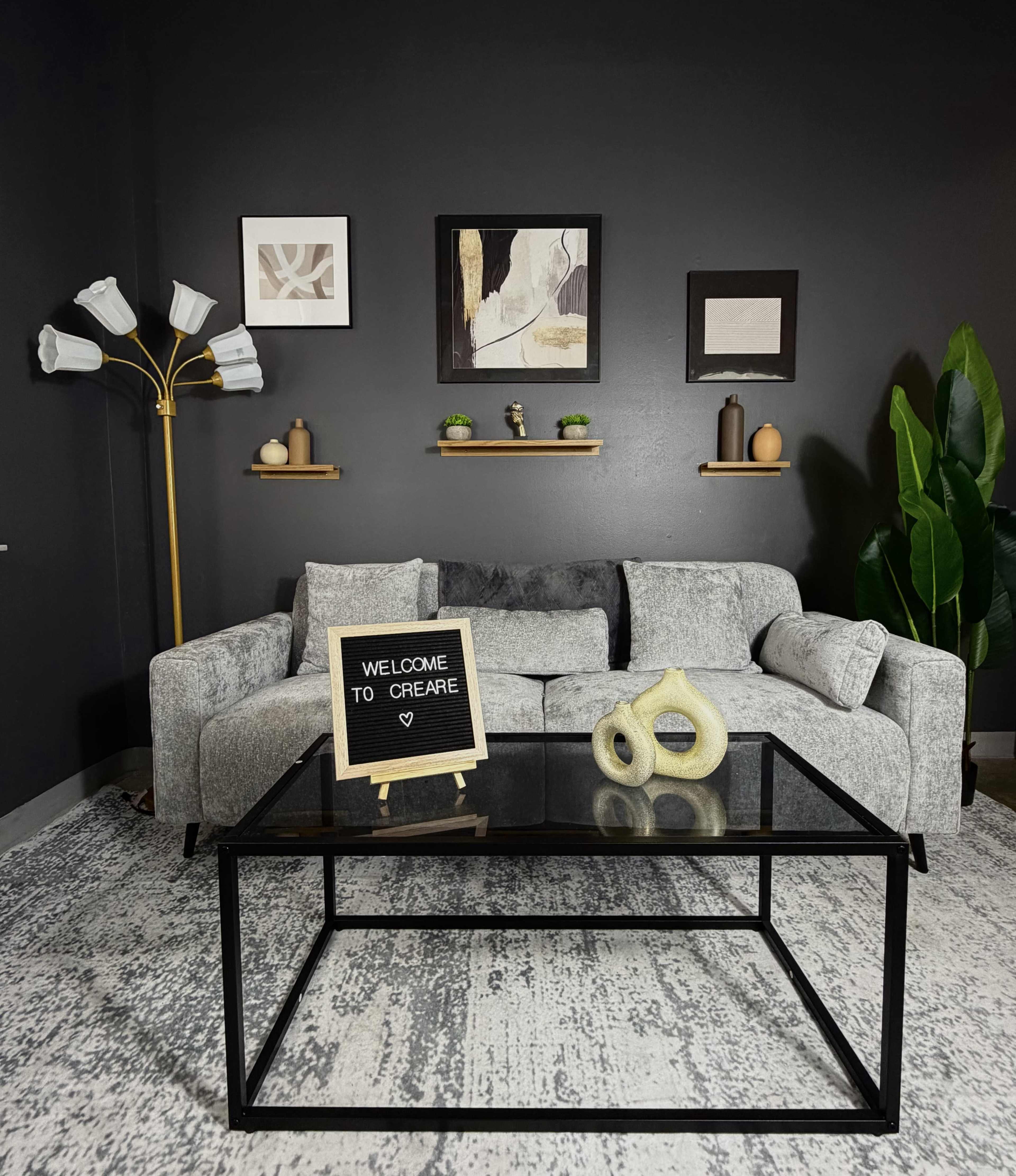 The image shows a modern living room with a gray sofa, a glass coffee table, and decorative wall art, complemented by plants and a floor lamp.