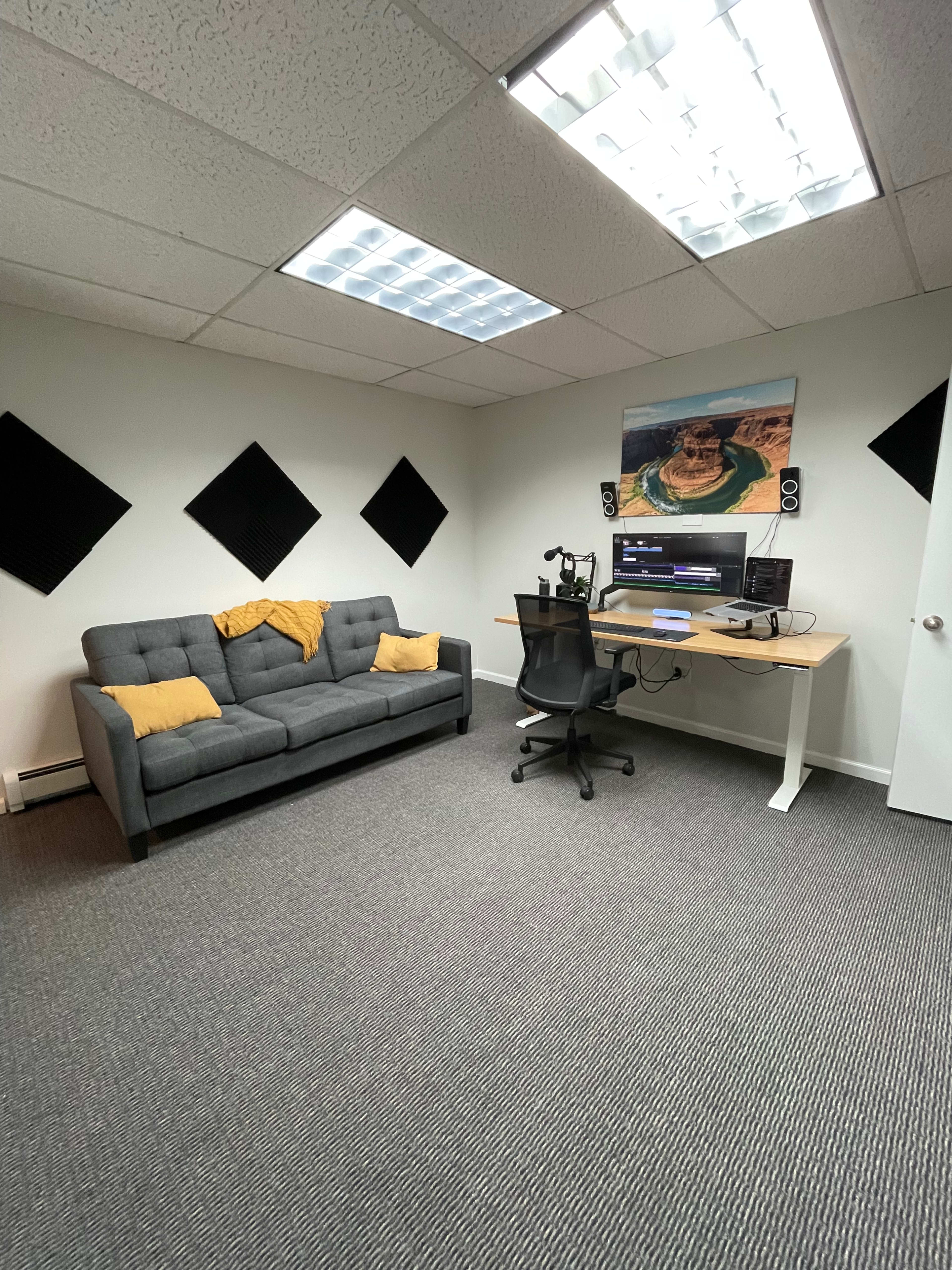 A small office space featuring a gray sofa with yellow throw pillows, a desk with audio equipment, and soundproofing panels on the walls.