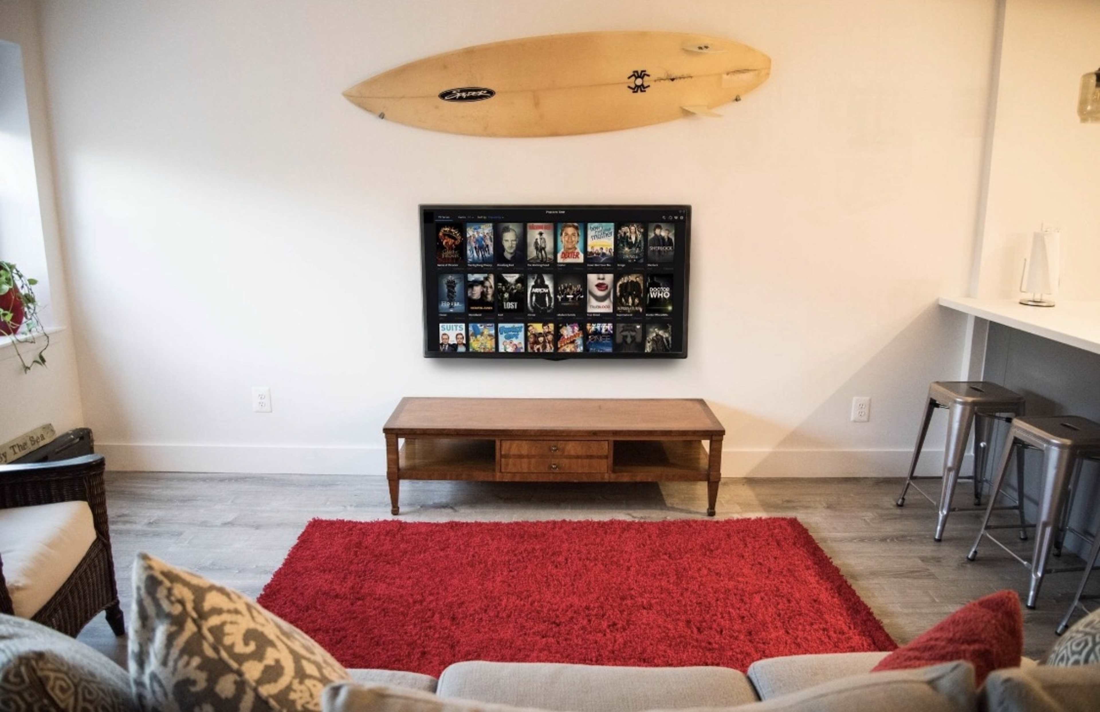 A living room features a wooden coffee table in front of a wall-mounted TV displaying a collection of movie covers, with a surfboard hanging above it and a red area rug on the floor.