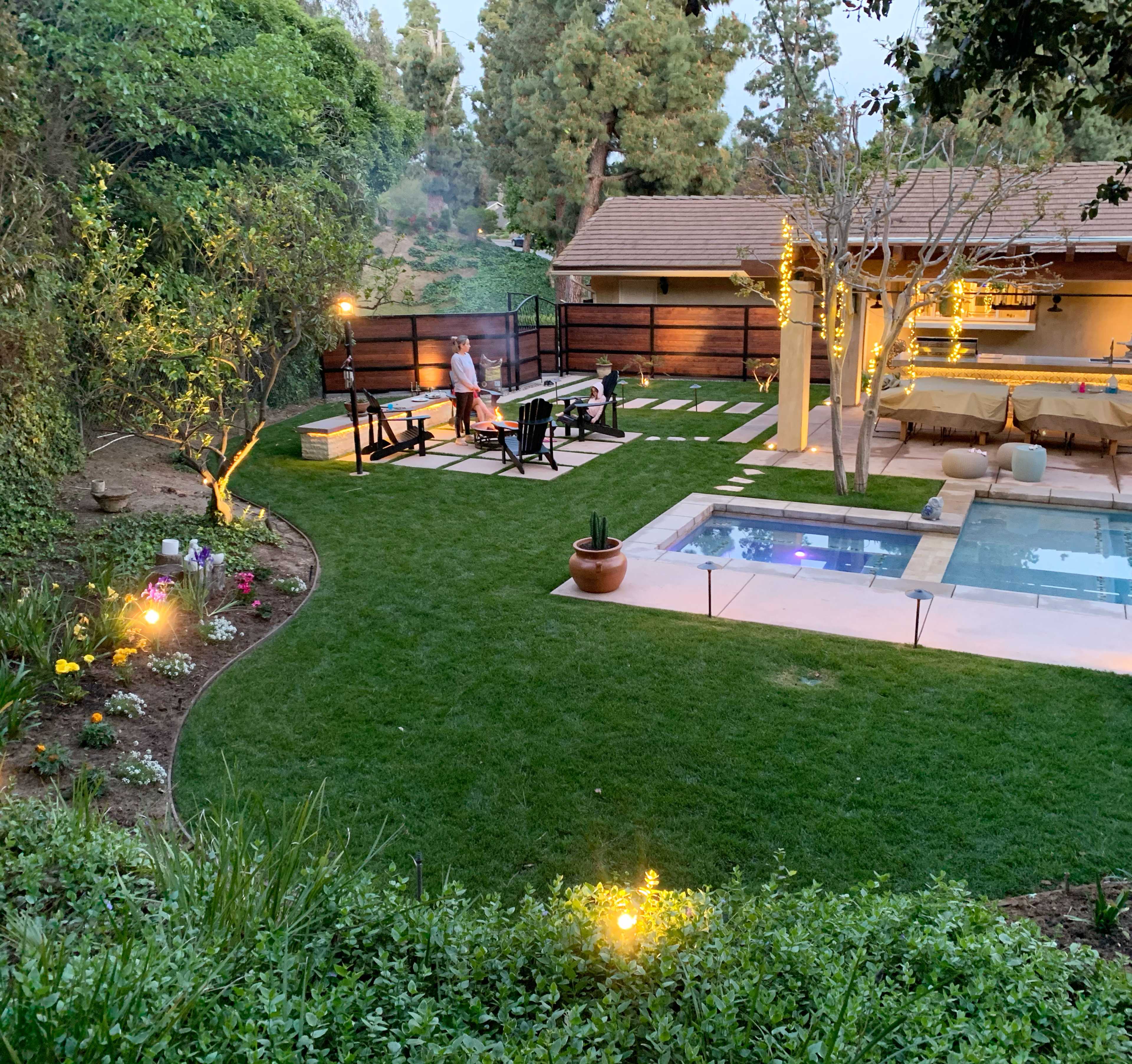 A landscaped backyard features a seating area around a fire pit, a swimming pool, and illuminated pathways amidst greenery and blooming flowers.