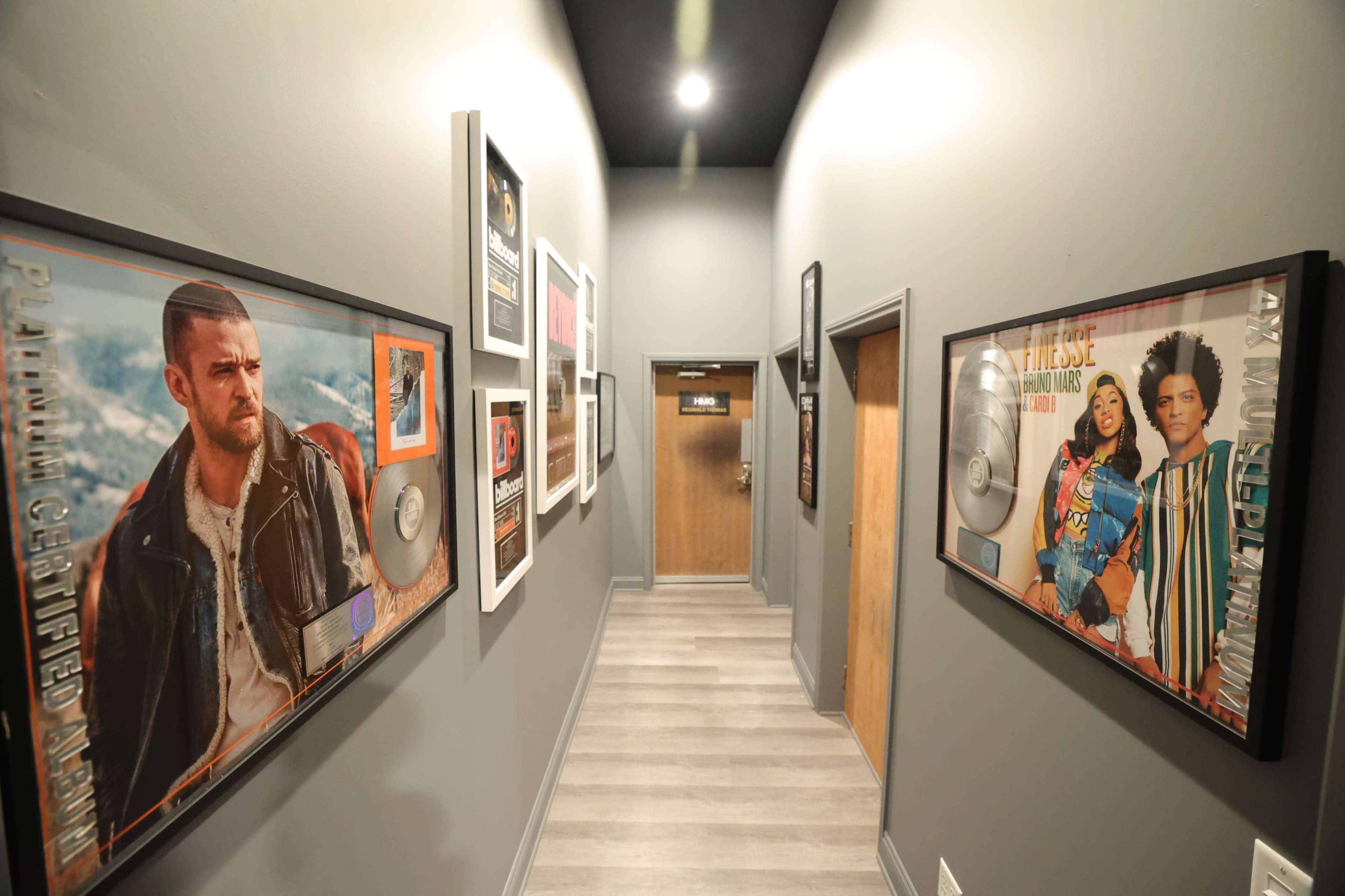 The image depicts a hallway lined with framed music awards and posters of artists on the walls, leading to a wooden door at the far end.