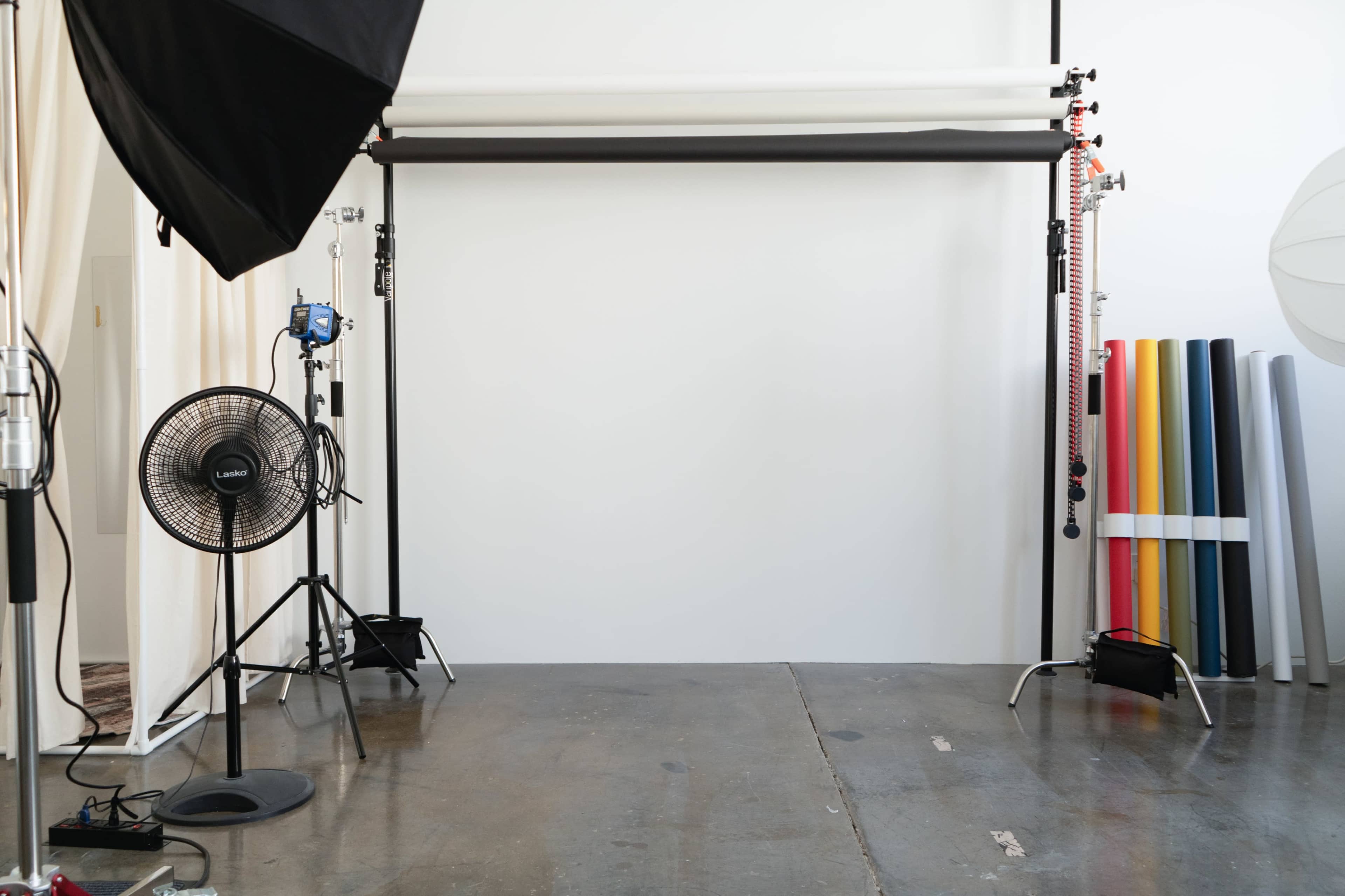 The image shows a photography studio with a white backdrop, lighting equipment, a fan, and rolls of colored paper.