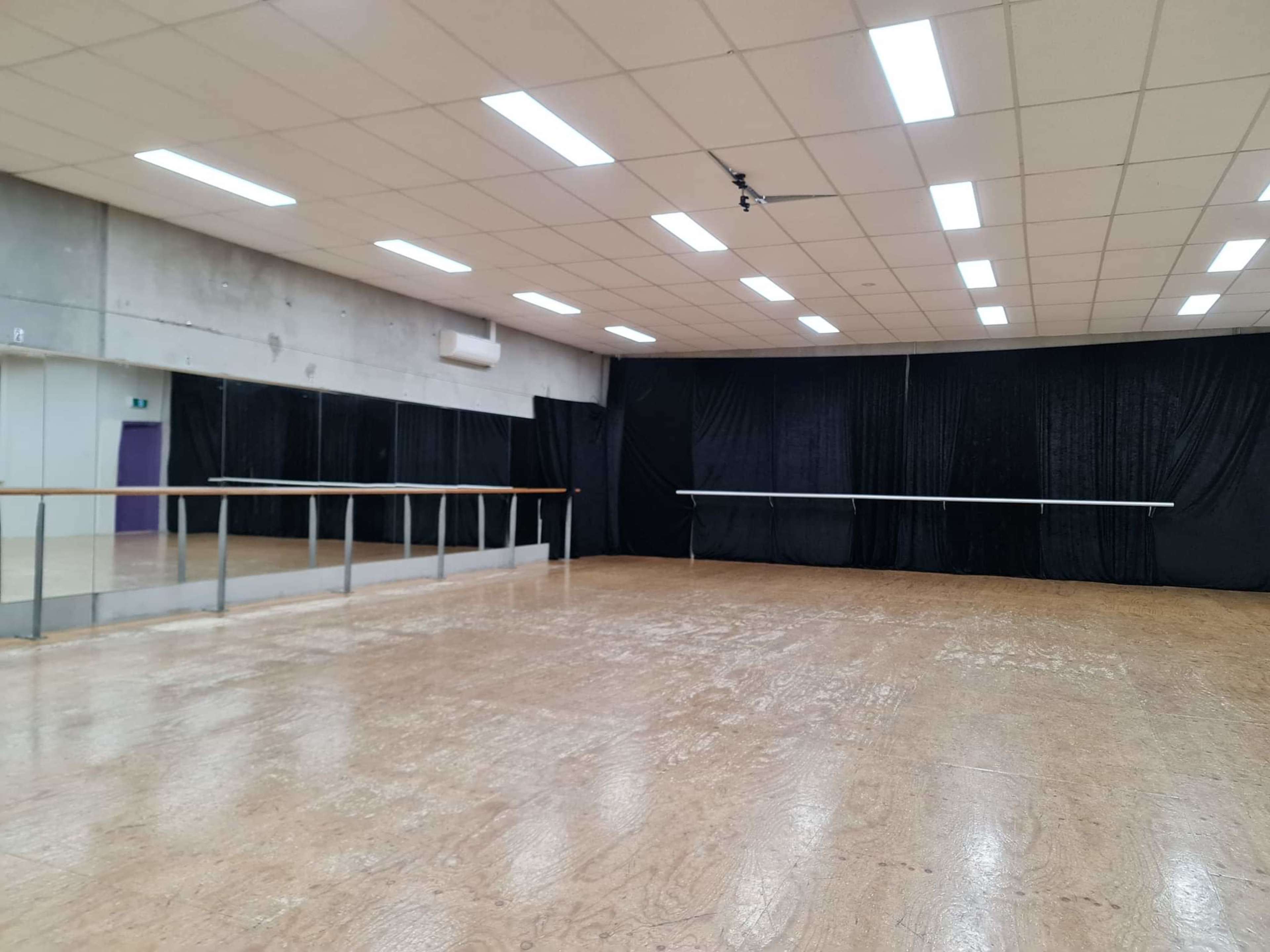 A spacious dance studio with a wooden floor, mirrored walls, and a black curtain at the far end.