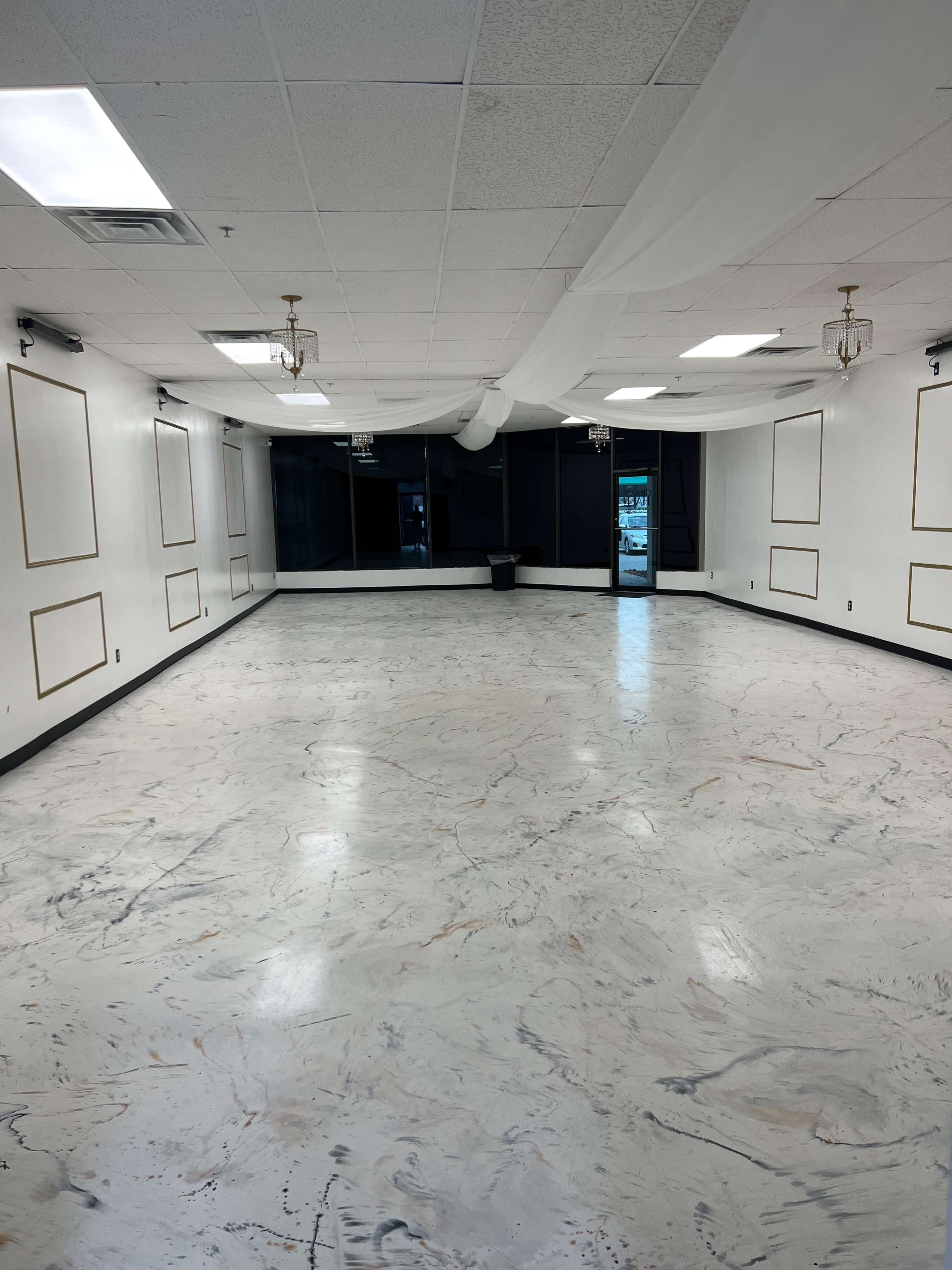 The image shows a spacious, empty room with a marbled floor, white walls, and decorative wall frames.